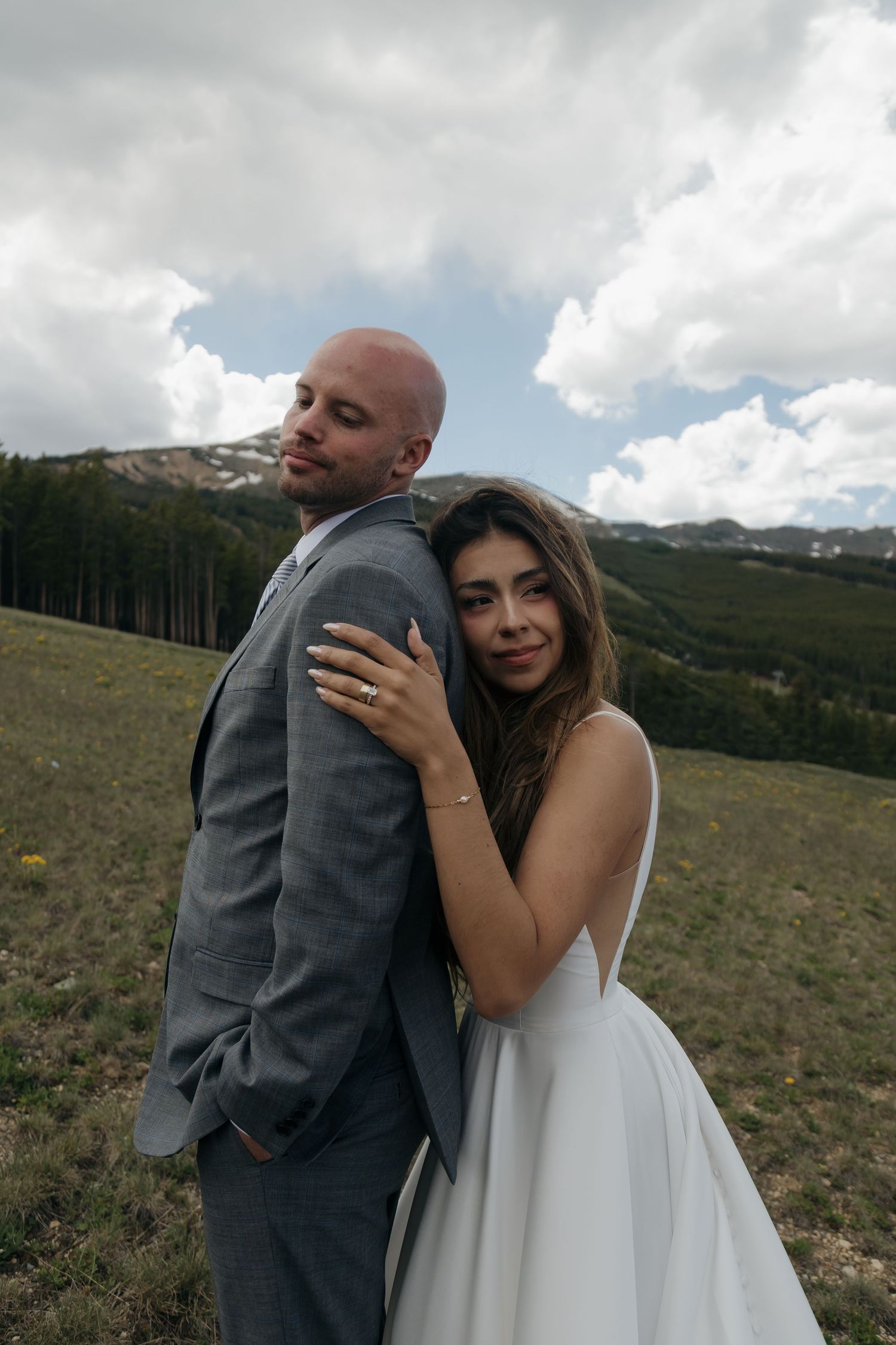 Bride leans against groom's back in a scenic mountain meadow with dramatic cloudy skies in the background.