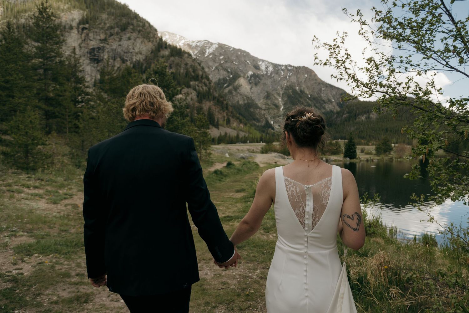 Bride and groom hold hands walking toward mountain lake landscape in elegant wedding attire.