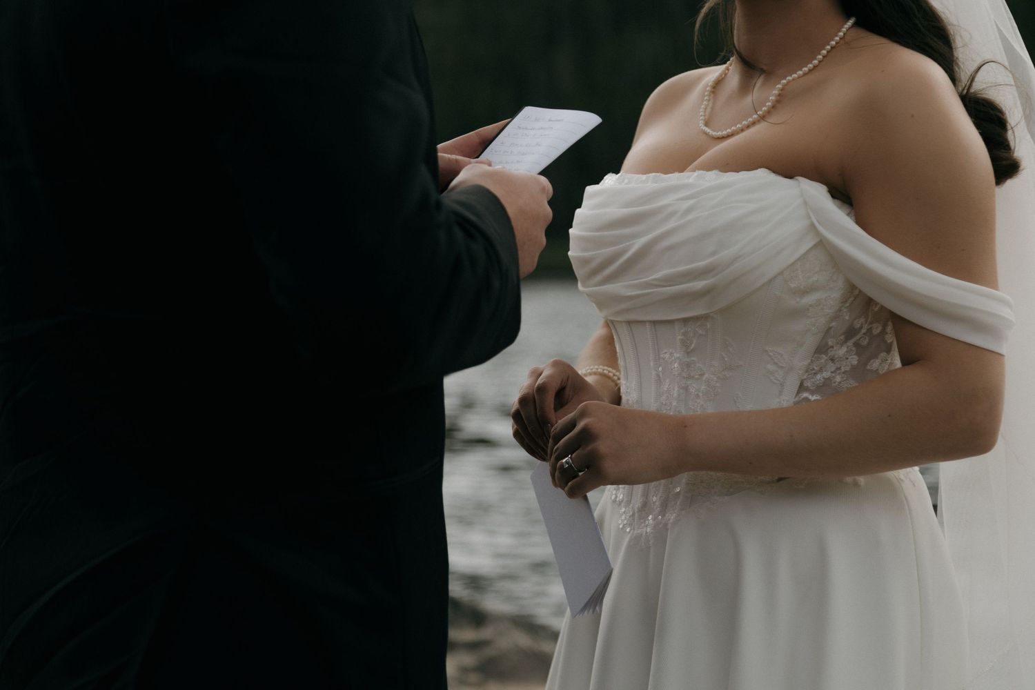 Bride in white off-shoulder wedding gown with pearl necklace listens as groom reads vows from a card outdoors.