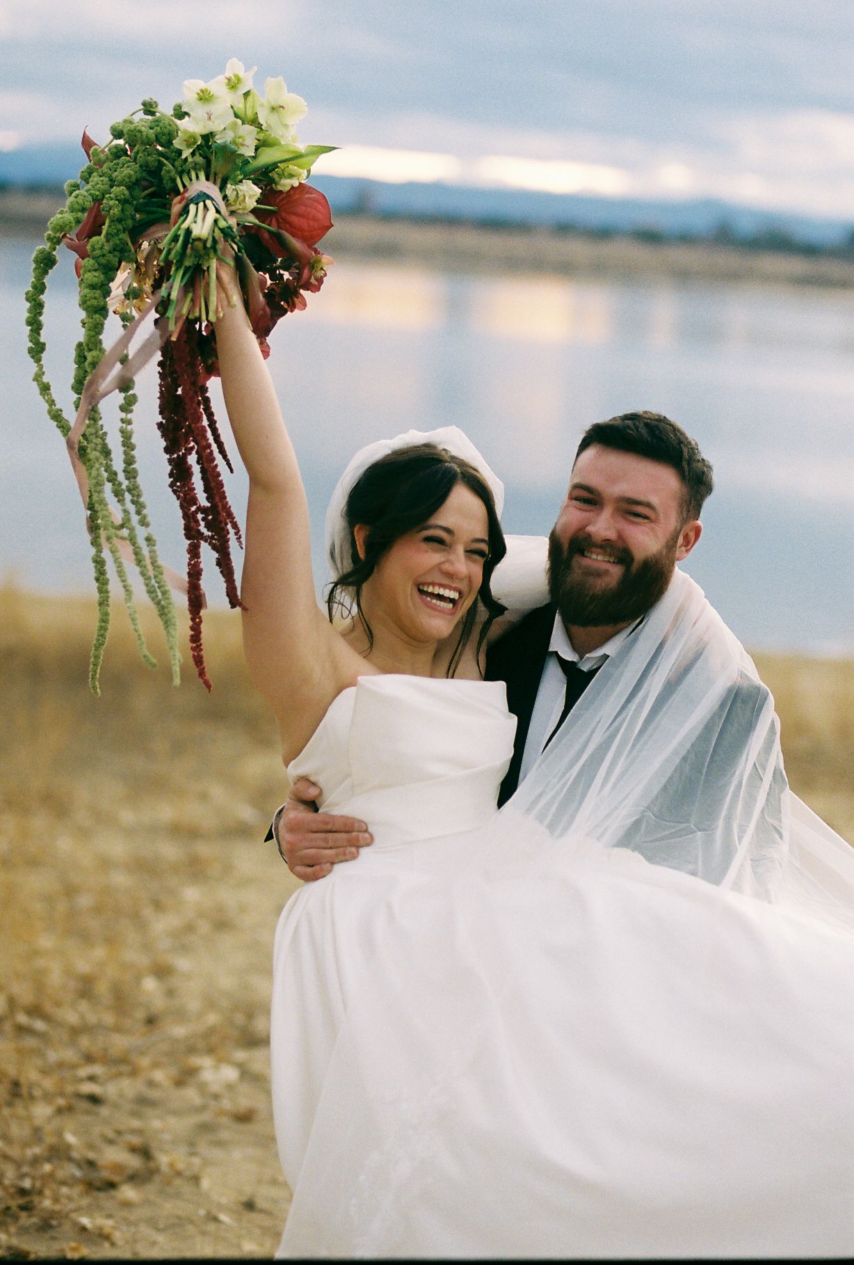 A joyful wedding portrait on a beach with a dramatic floral bouquet held high.