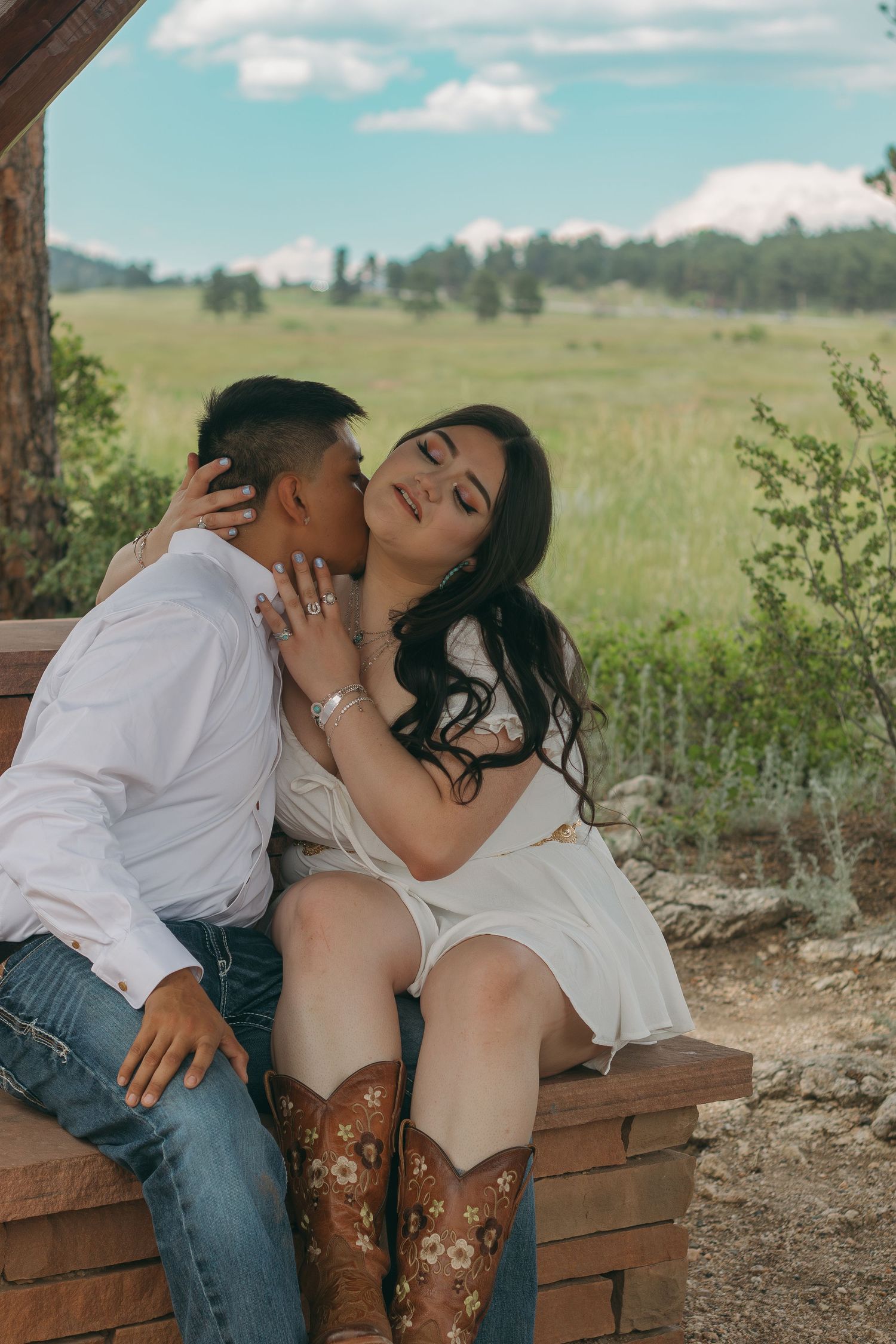 Intimate couple shares a tender moment on wooden bench with scenic mountain landscape behind them.