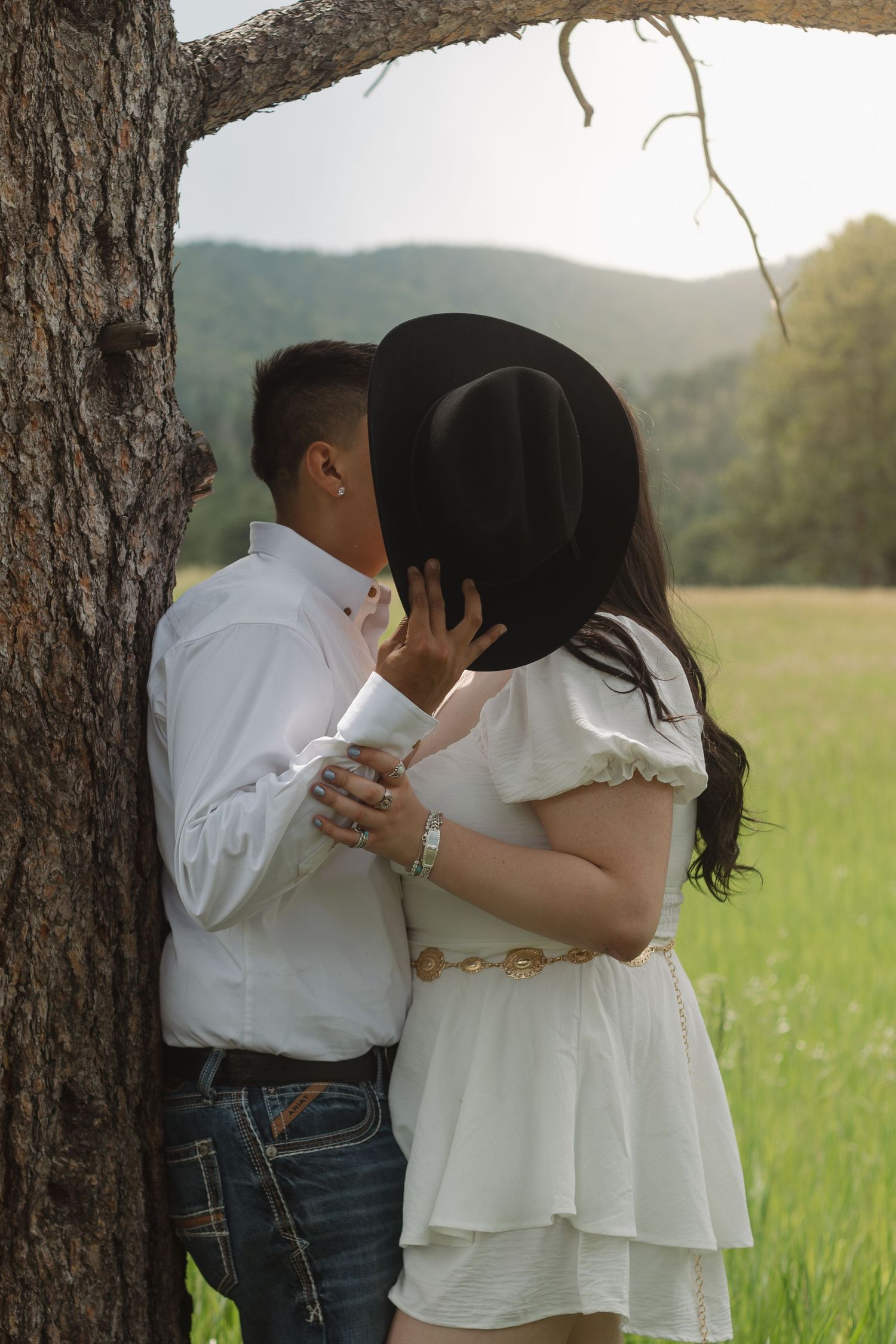 A couple shares a romantic moment behind a cowboy hat against a tree trunk.
