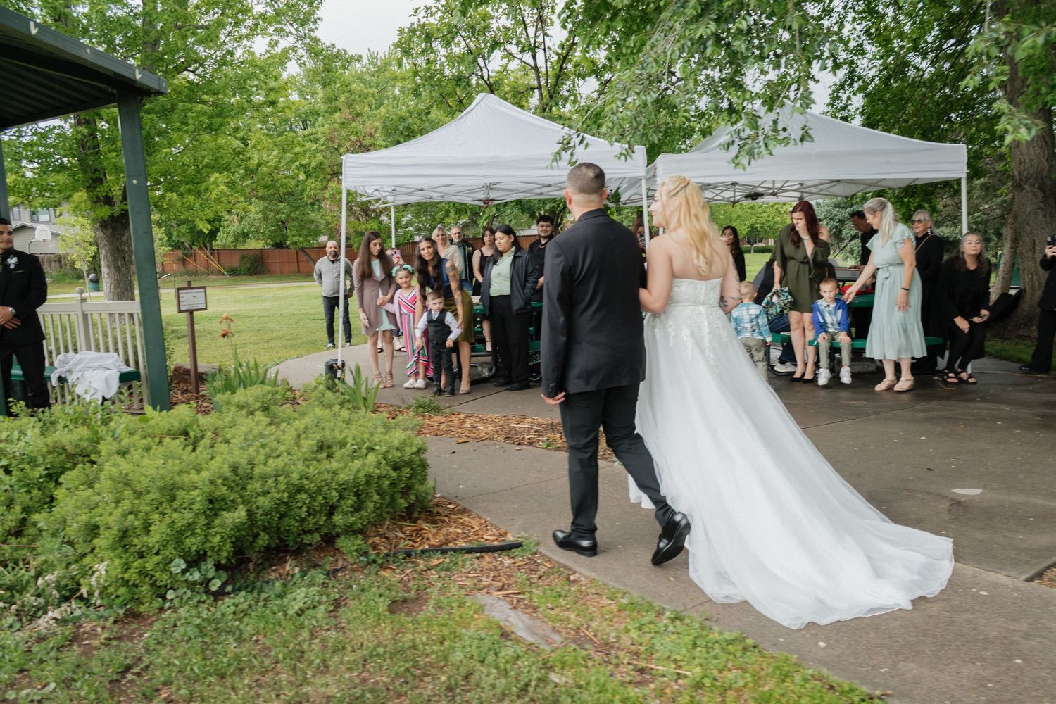 A couple walks down the aisle at an outdoor wedding with guests seated under white tents.