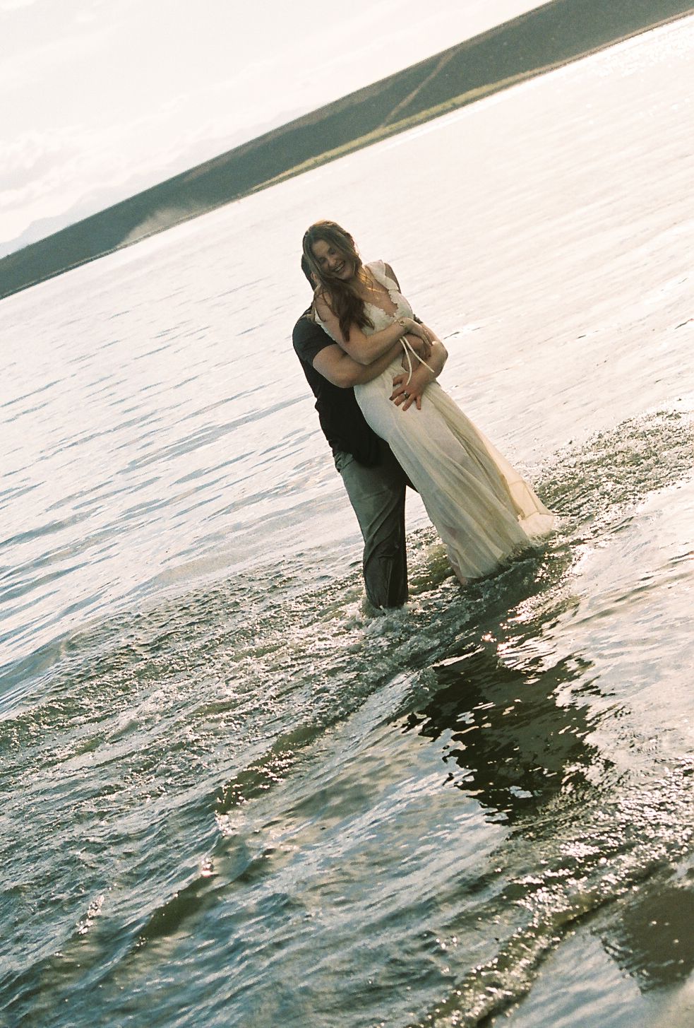 Couple embracing romantically while standing in shallow ocean water, bride in white gown and groom in dark attire.
