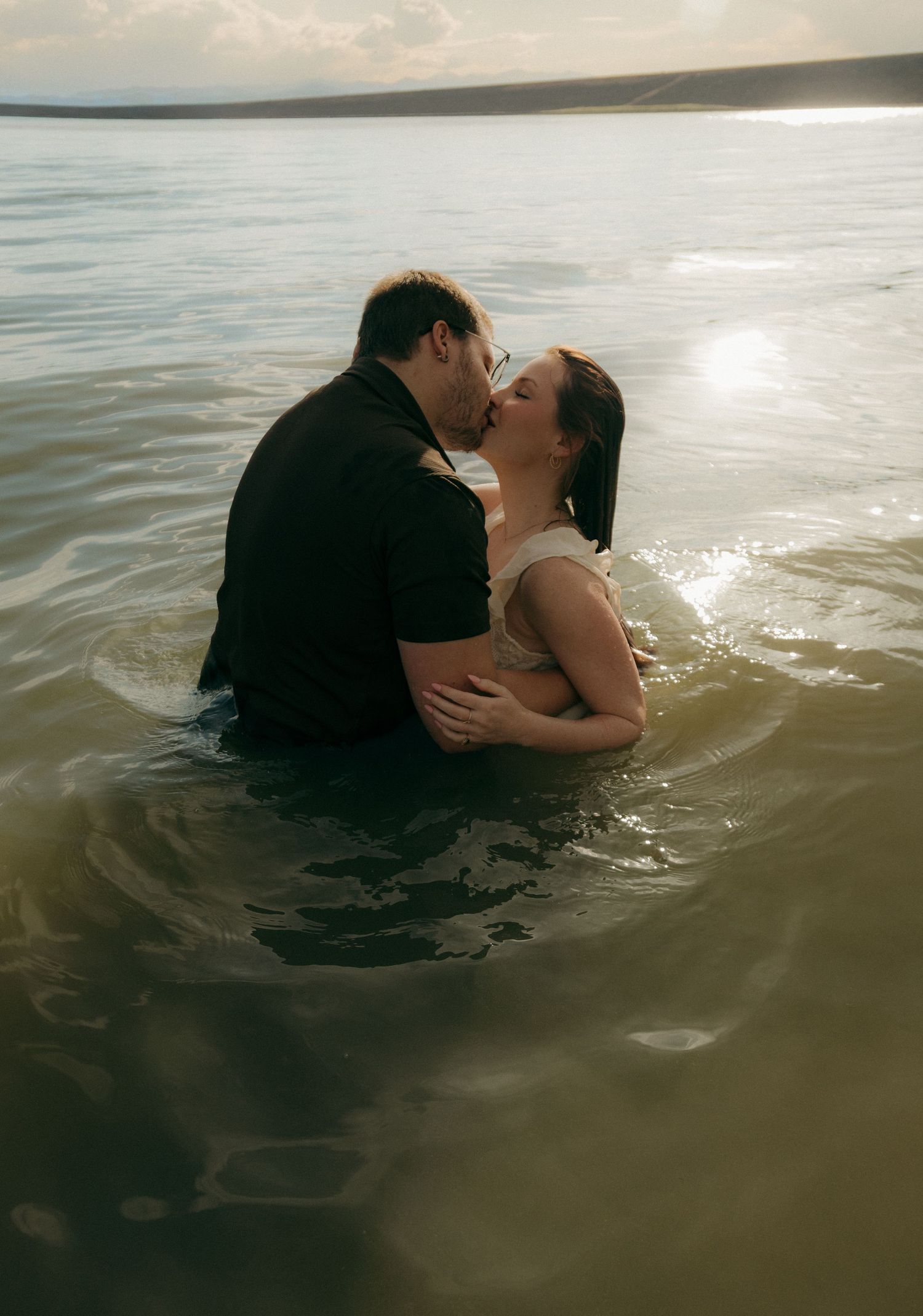 Couple sharing a passionate kiss while submerged waist-deep in a sunlit lake during a romantic engagement photo session.