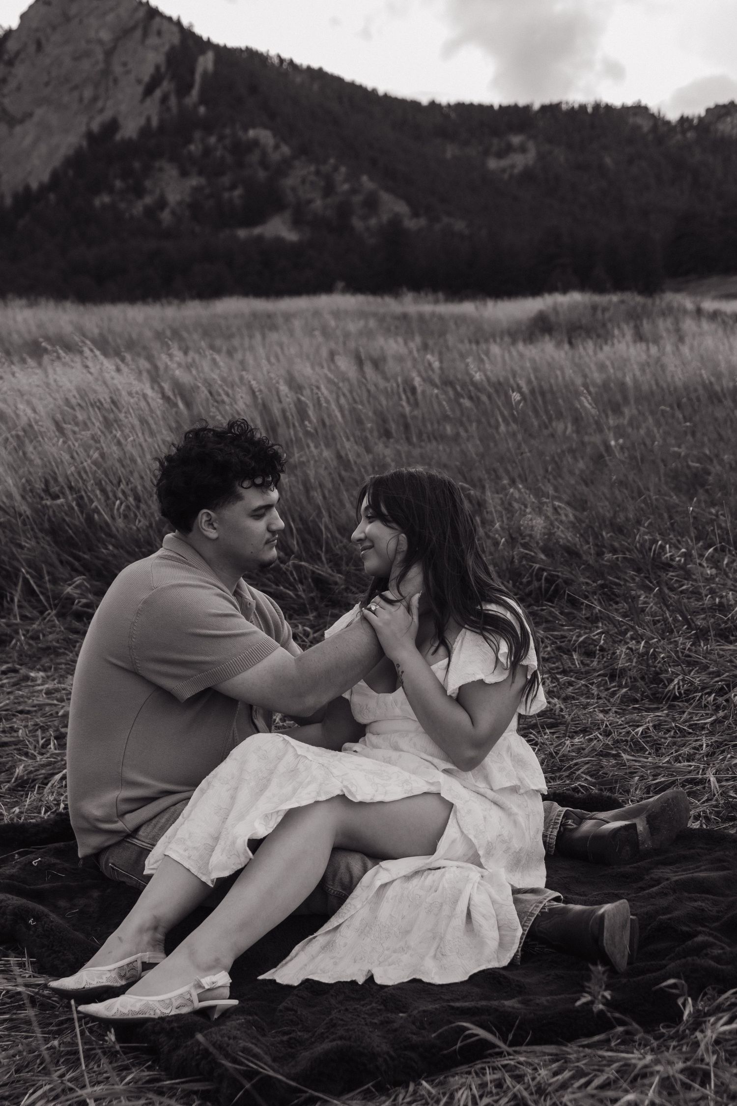 Black and white photo of a couple sitting in a field with mountains behind them.