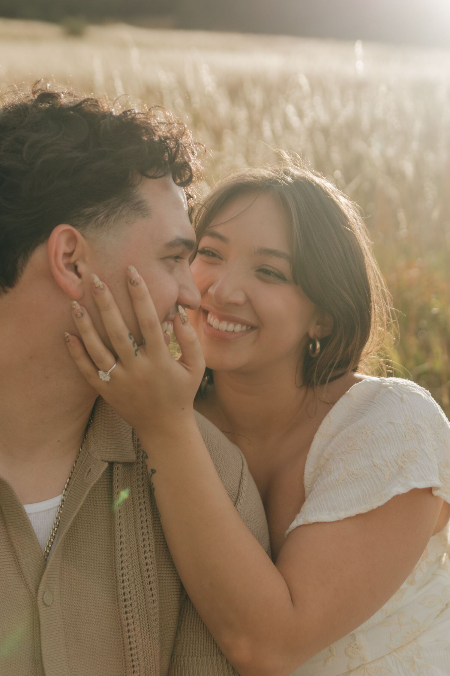 Smiling woman gently holds mans face as they share an intimate moment in a sun-drenched field.