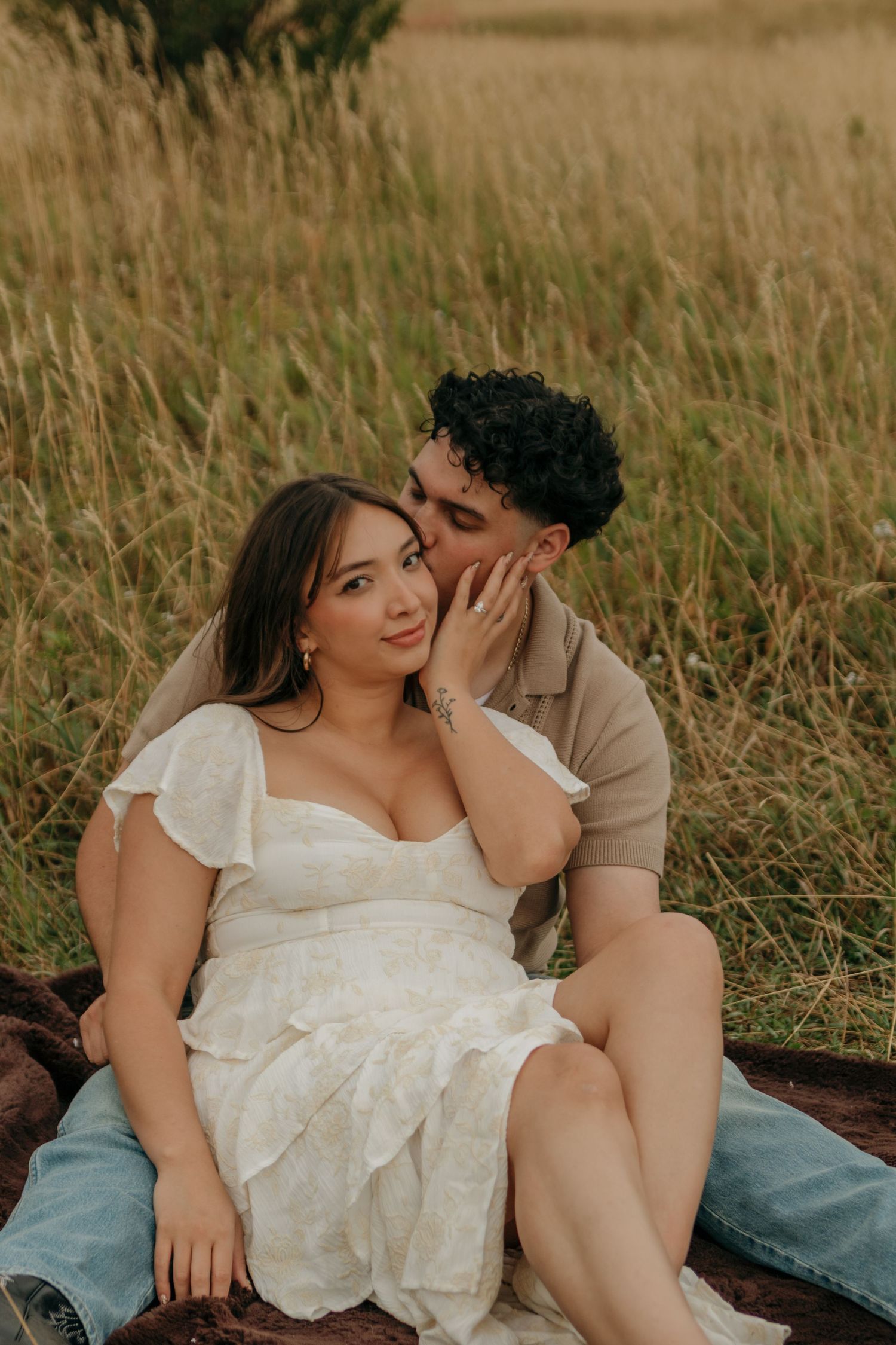 A man kisses a womans cheek as she smiles in a white dress while sitting in a grassy field.