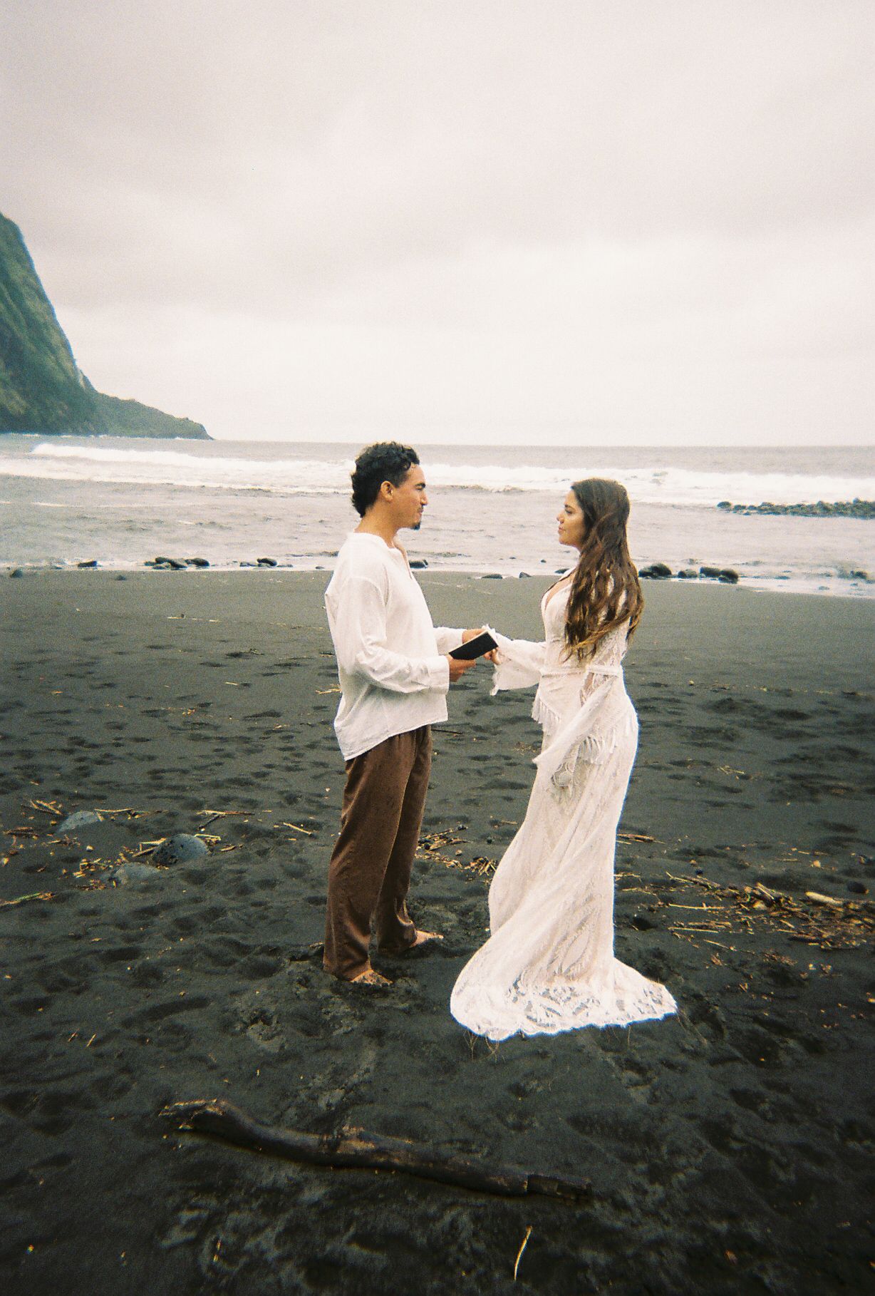 Couple exchanging vows on a black sand beach with ocean waves and misty cliffs, bride in lace gown.