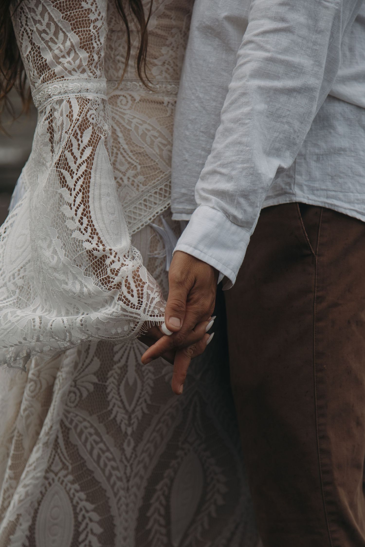 Close-up of interracial couple holding hands, bride wearing intricate lace boho wedding dress.