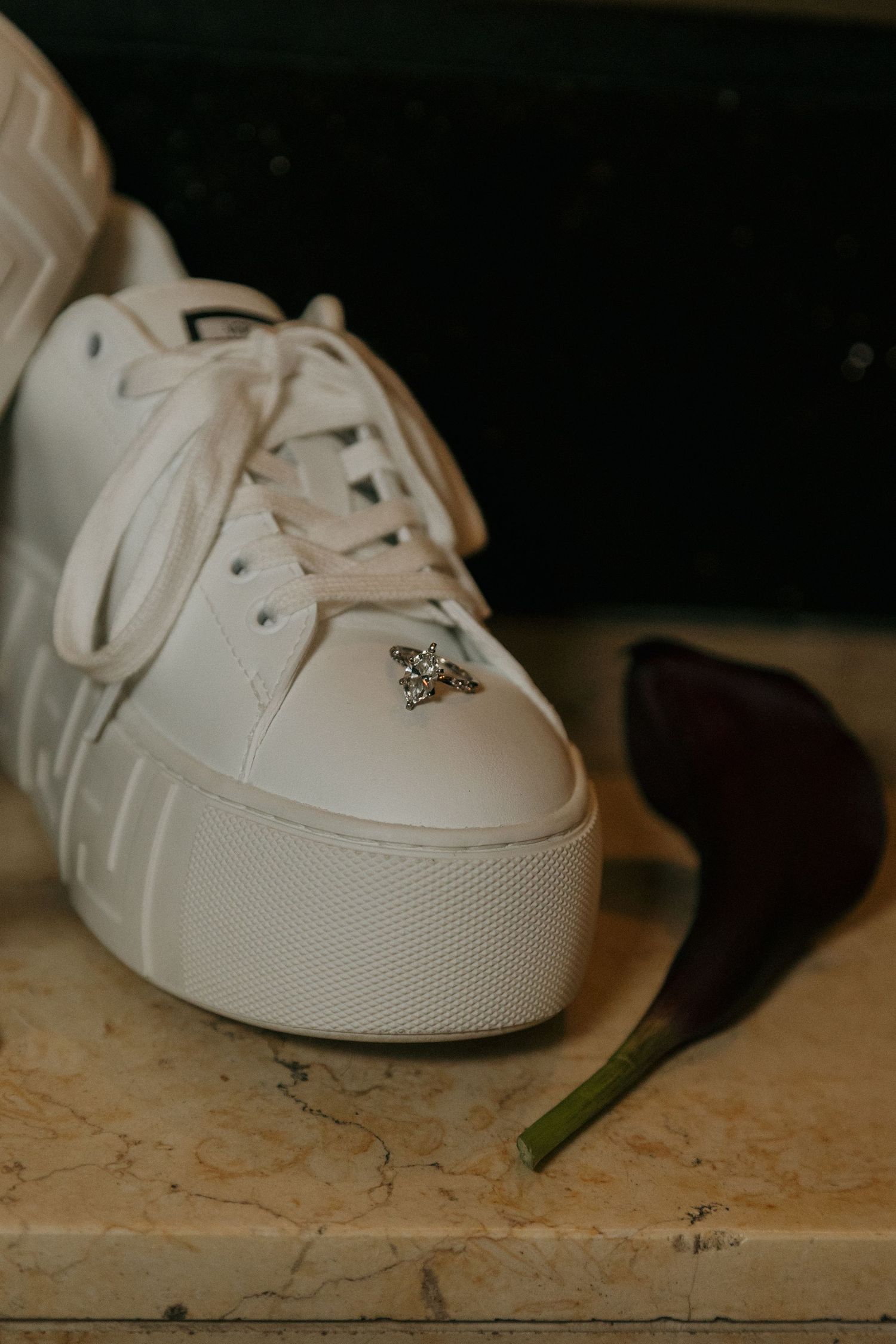 White platform sneaker with crystal embellishment on marble surface beside a dark flower petal.