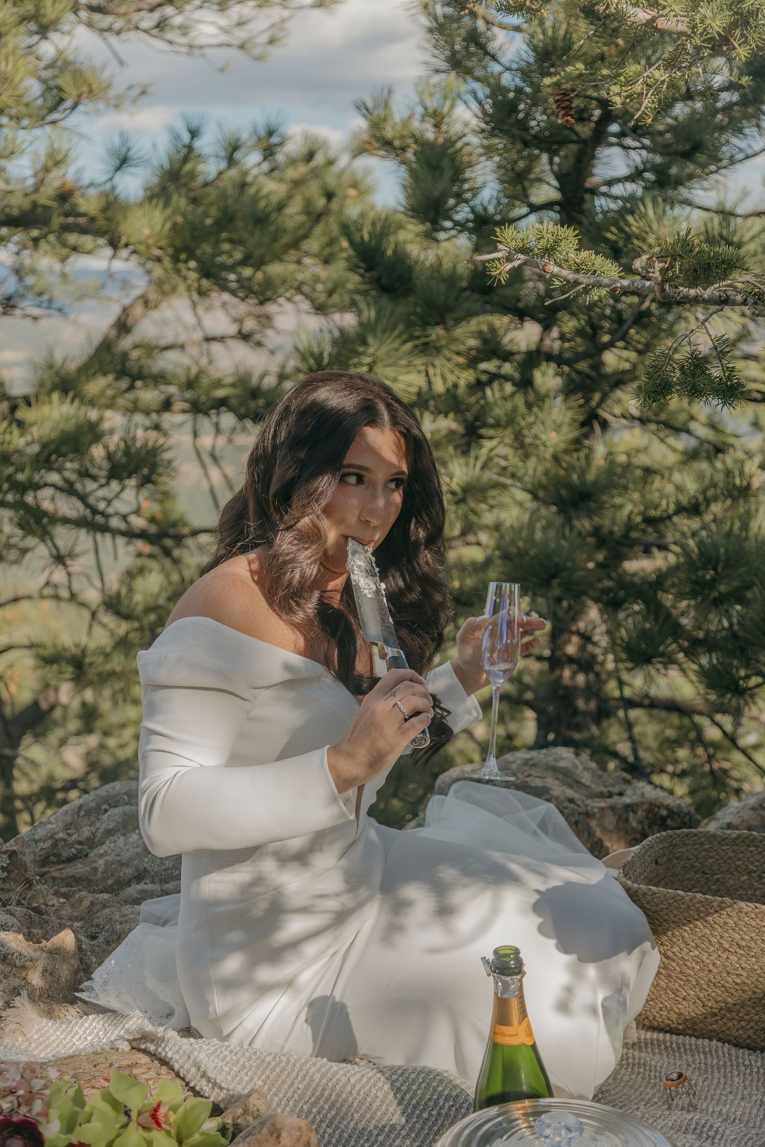 Bride in an off-shoulder white gown pours champagne while seated on rocks surrounded by pine trees outdoors.
