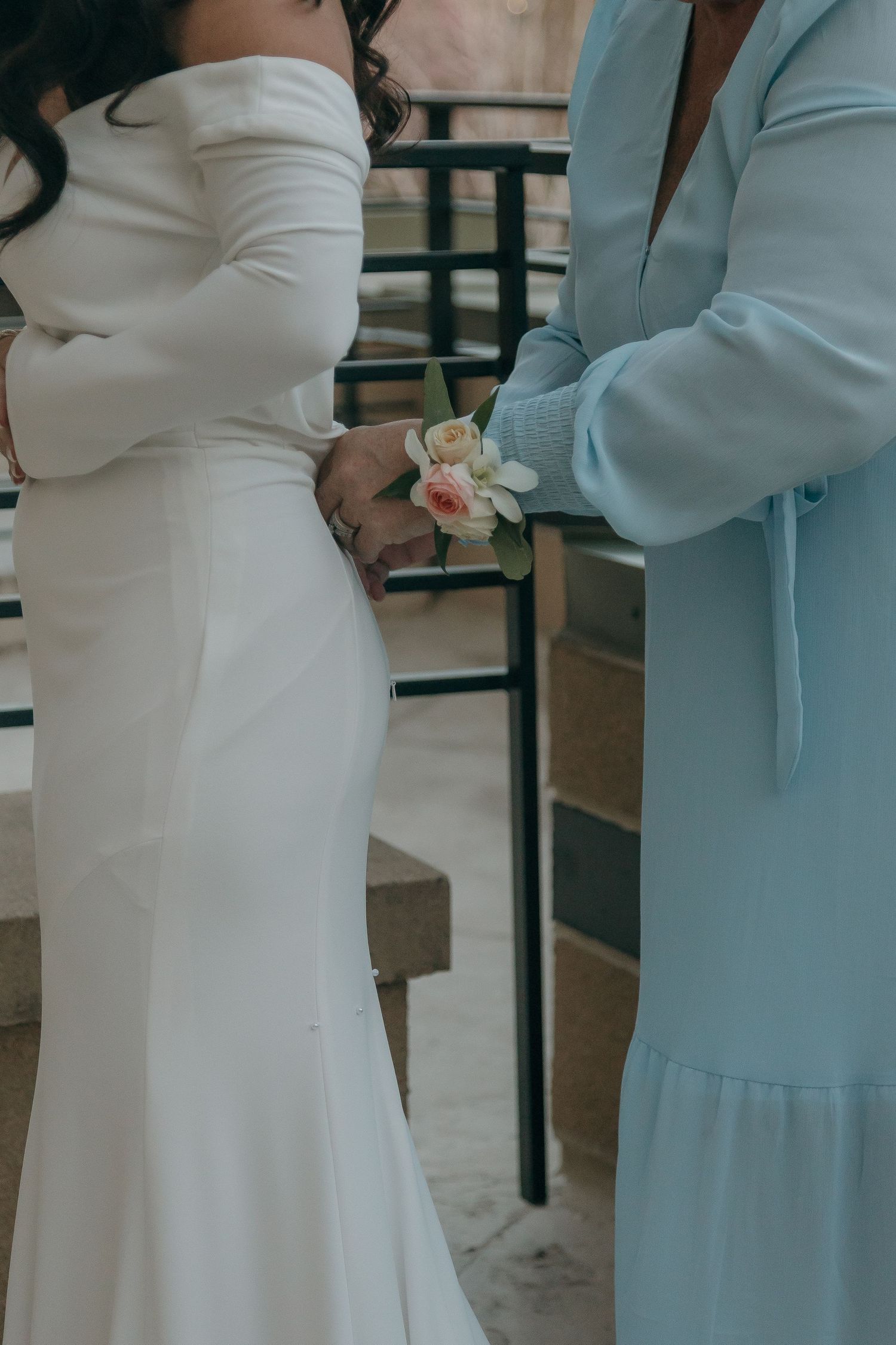 Bride in white off-shoulder gown receives floral corsage from guest in light blue dress.
