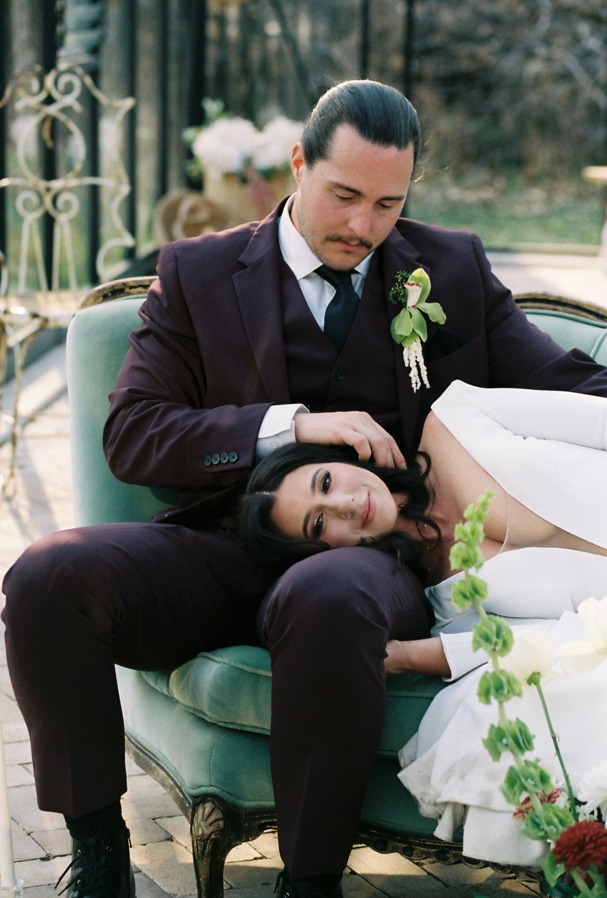 Bride resting head on groom's lap as they sit together on vintage teal velvet chair outdoors.