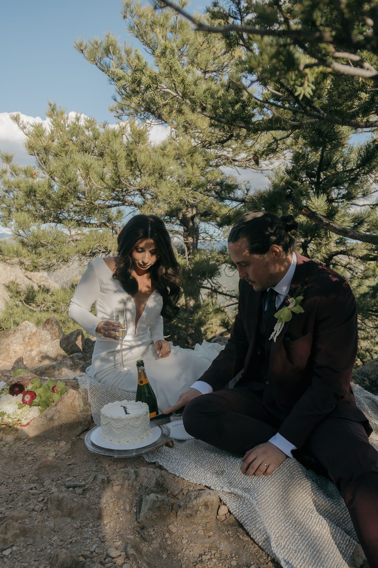 Bride and groom cutting white wedding cake outdoors on rocky mountain ledge with champagne bottle beside them.