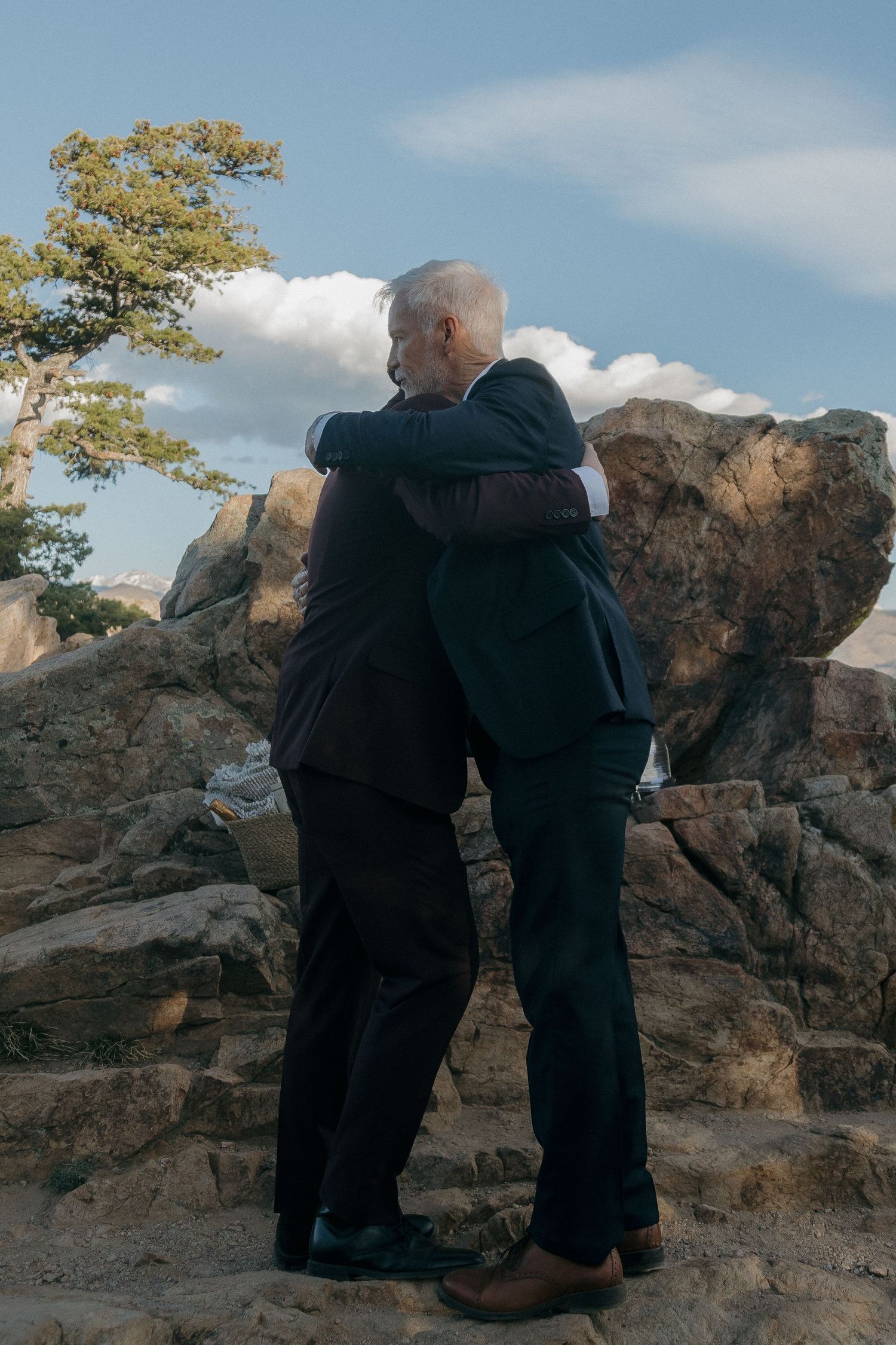 Two men in dark suits sharing a heartfelt hug on a rocky outdoor wedding venue.