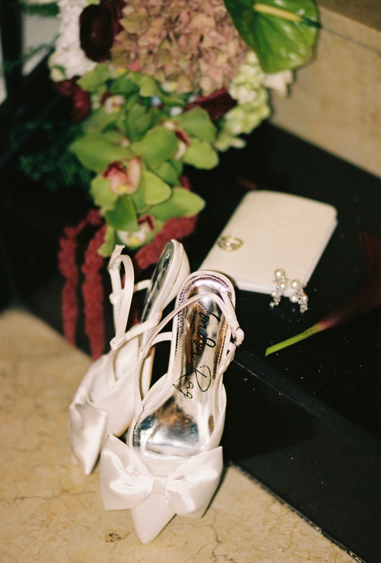 White satin bow wedding heels displayed beside floral bouquet and pearl earrings on dark surface.
