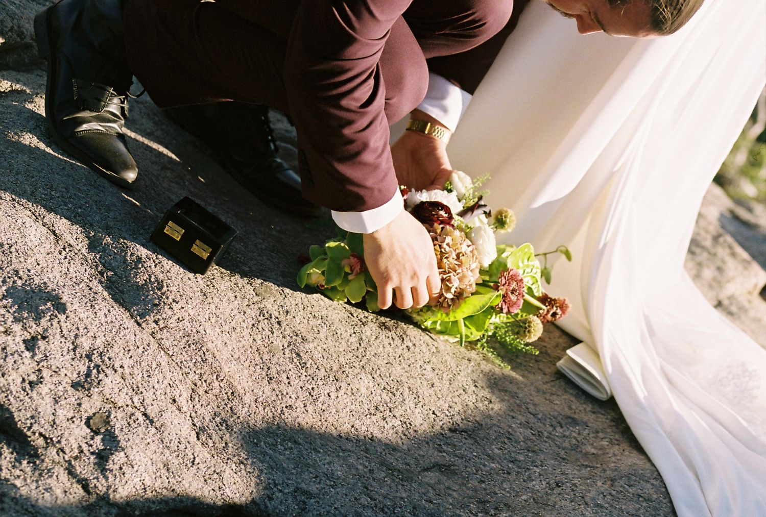 Groom in burgundy suit placing floral wreath on rocky ground beside bride in white dress.