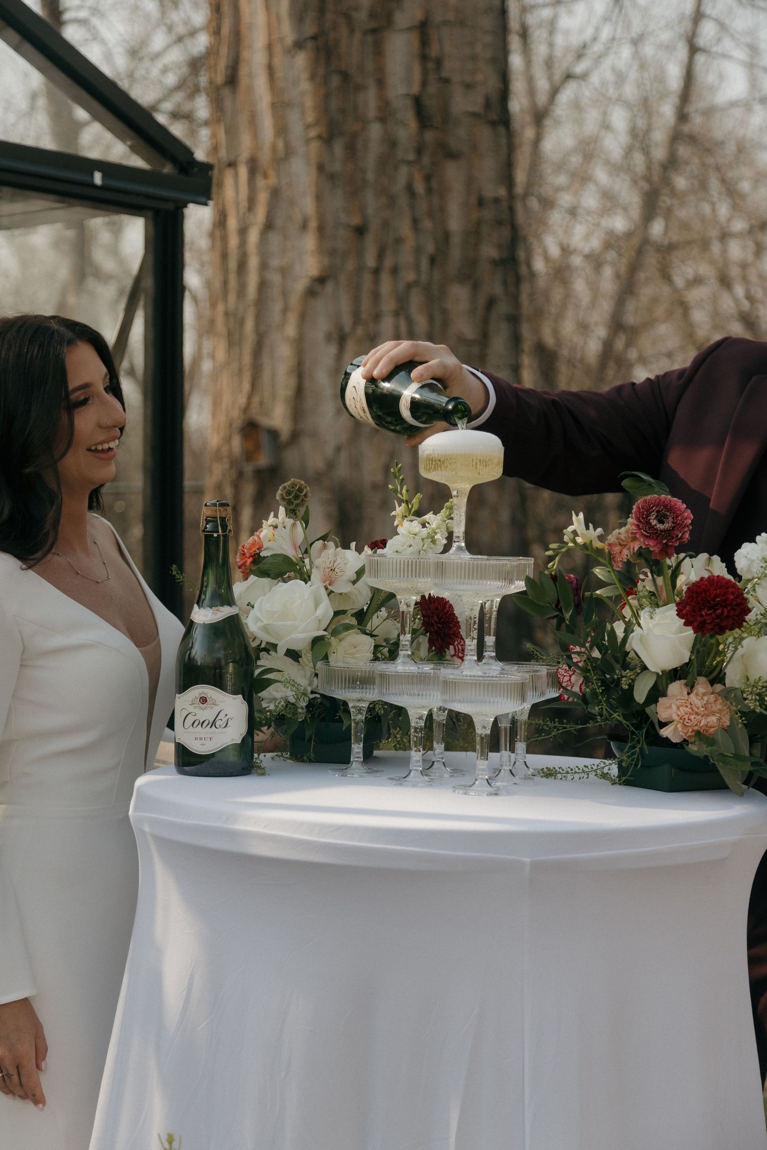 Bride smiles as groom pours champagne into a tower of glasses surrounded by white and red florals.