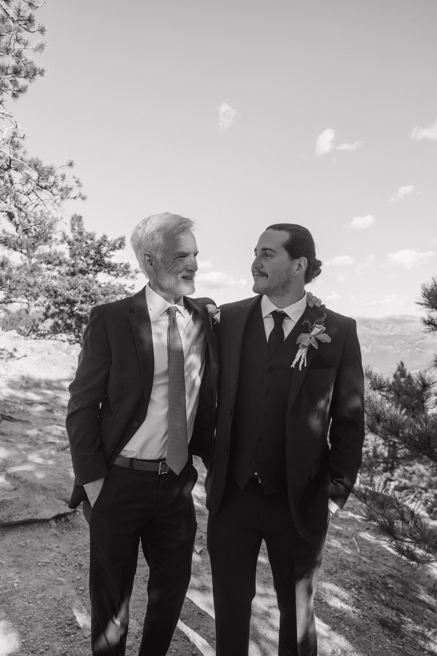Black and white portrait of groom and older man in suits smiling together at outdoor mountain wedding.