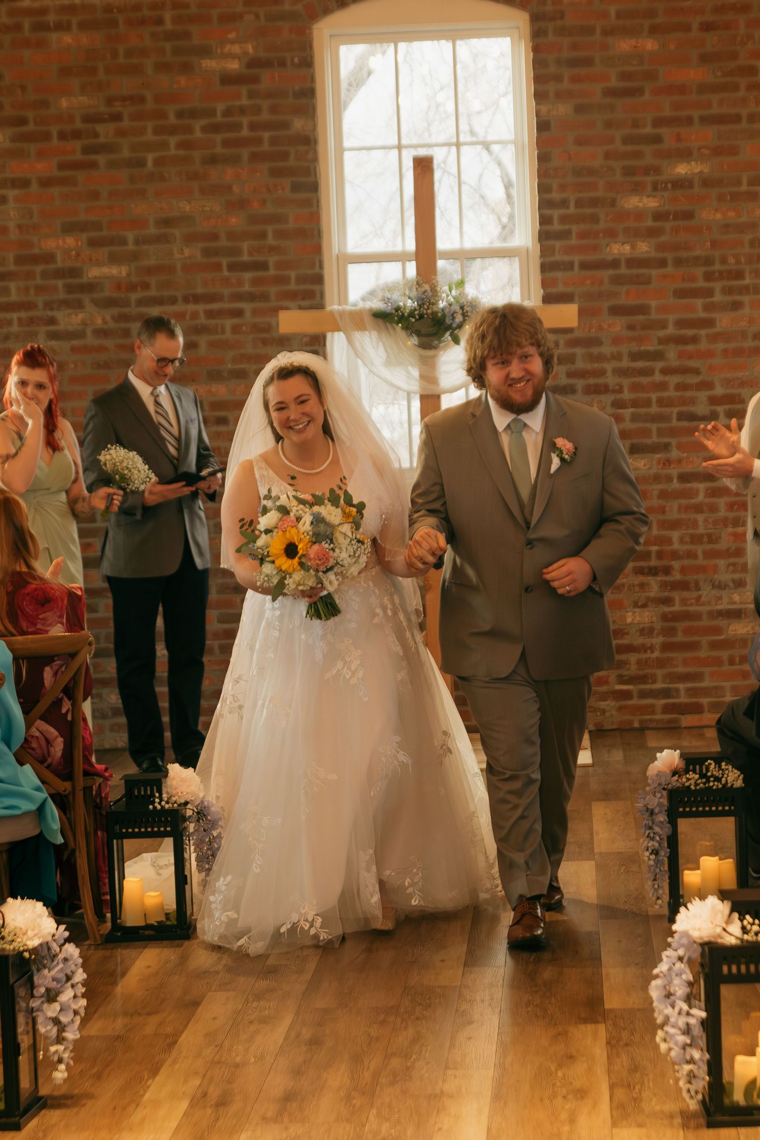Wedding couple walks down aisle decorated with lanterns in rustic brick venue with natural light streaming through windows.