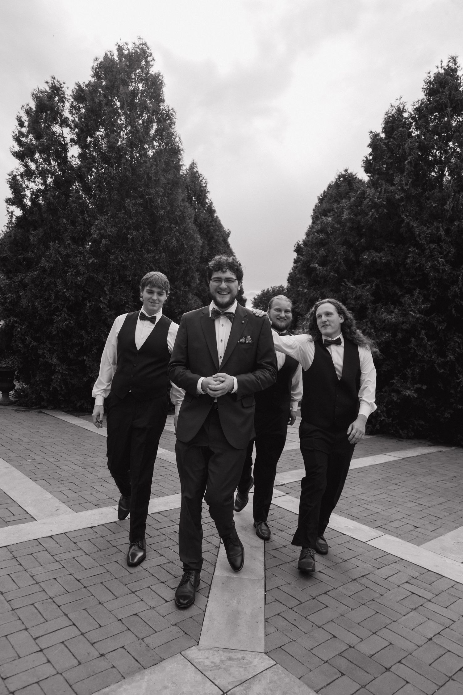 Four groomsmen in black tuxedos and bow ties walking confidently down a brick pathway lined with tall trees.