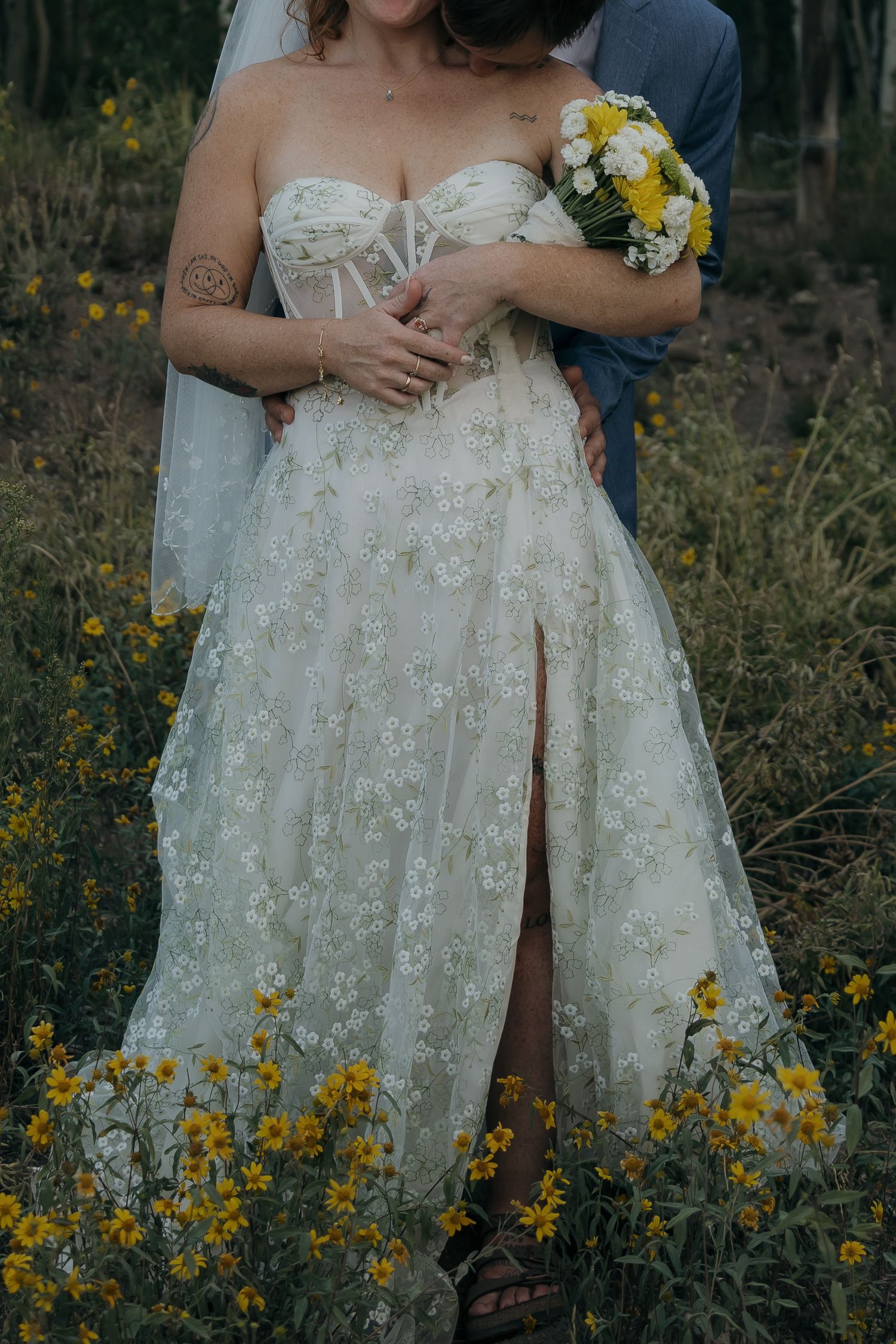 Bride in floral lace wedding dress with slit holding yellow and white bouquet in wildflower field.