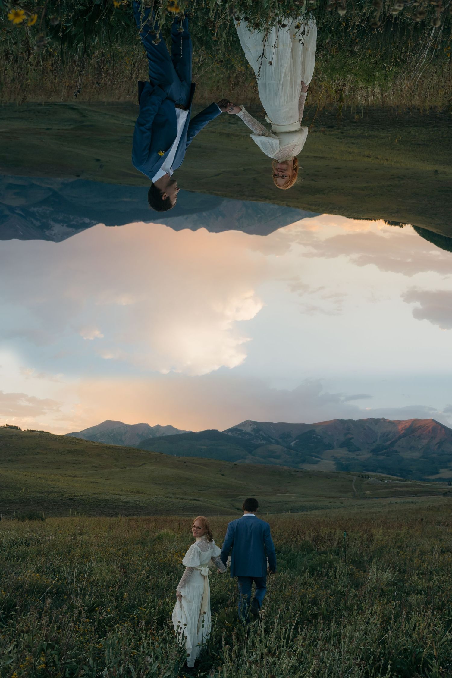 Creative mirror reflection photo of bride and groom holding hands walking through mountain meadow at golden hour.