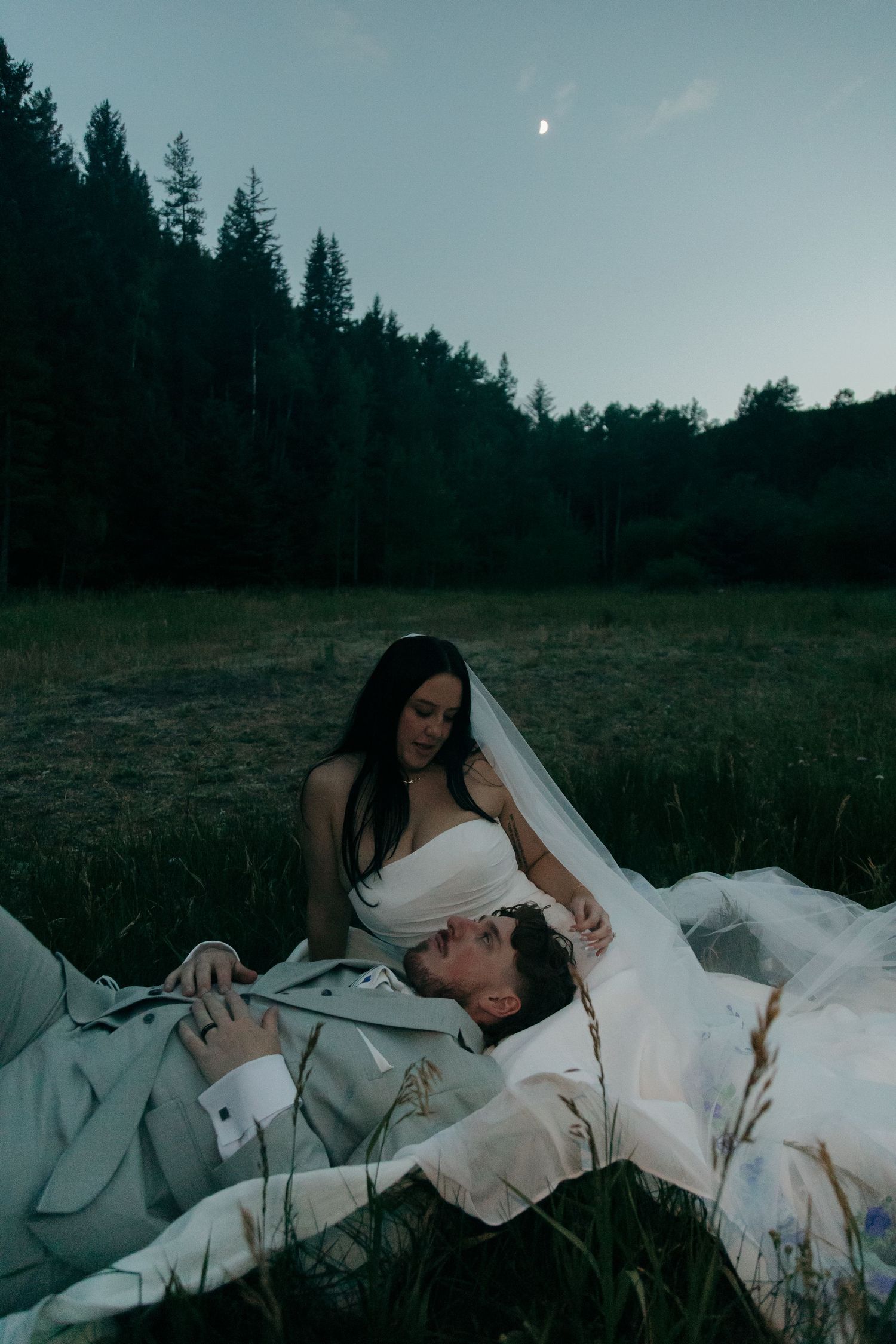 Bride and groom rest in moonlit meadow, groom lying in bride's lap at dusk.