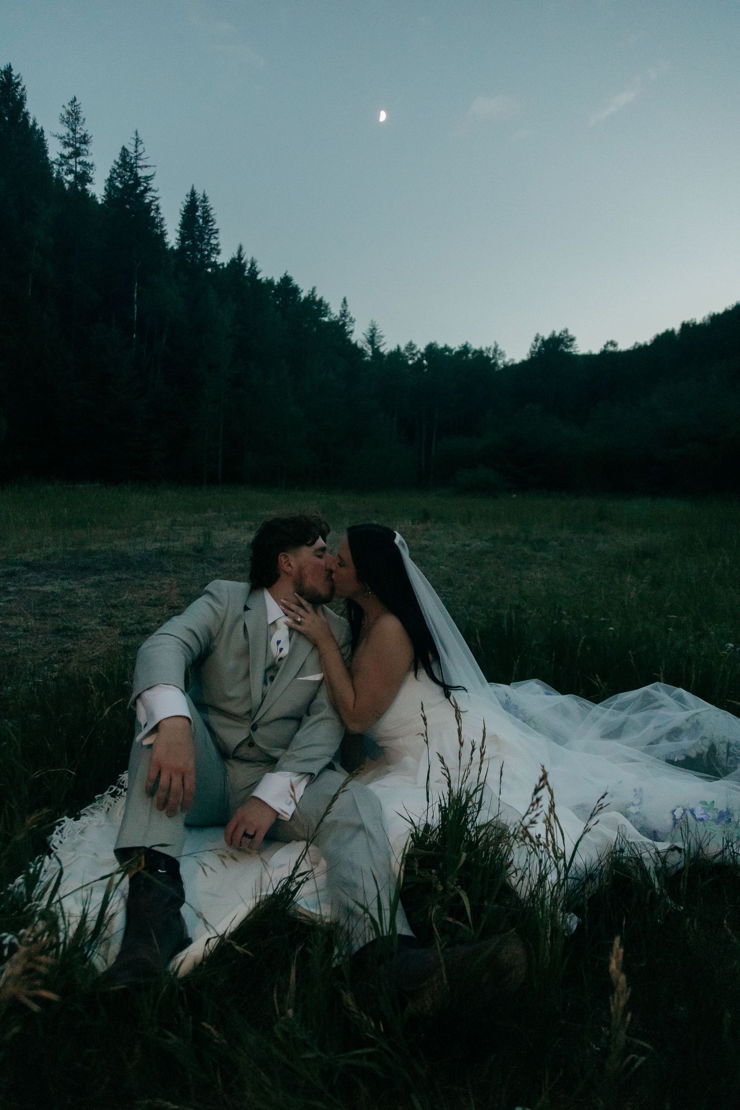 Newlyweds share a romantic kiss sitting in a moonlit meadow at dusk on their wedding night.