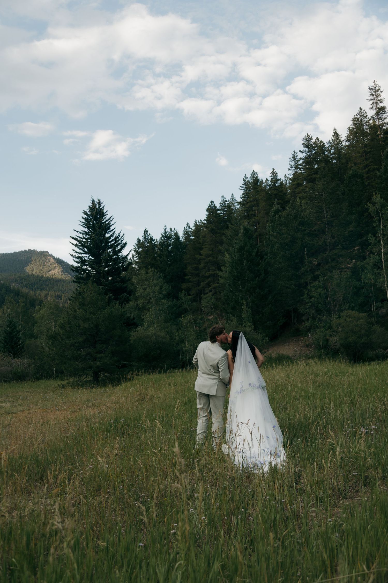 Bride and groom share intimate moment in green mountain meadow under cloudy sky.
