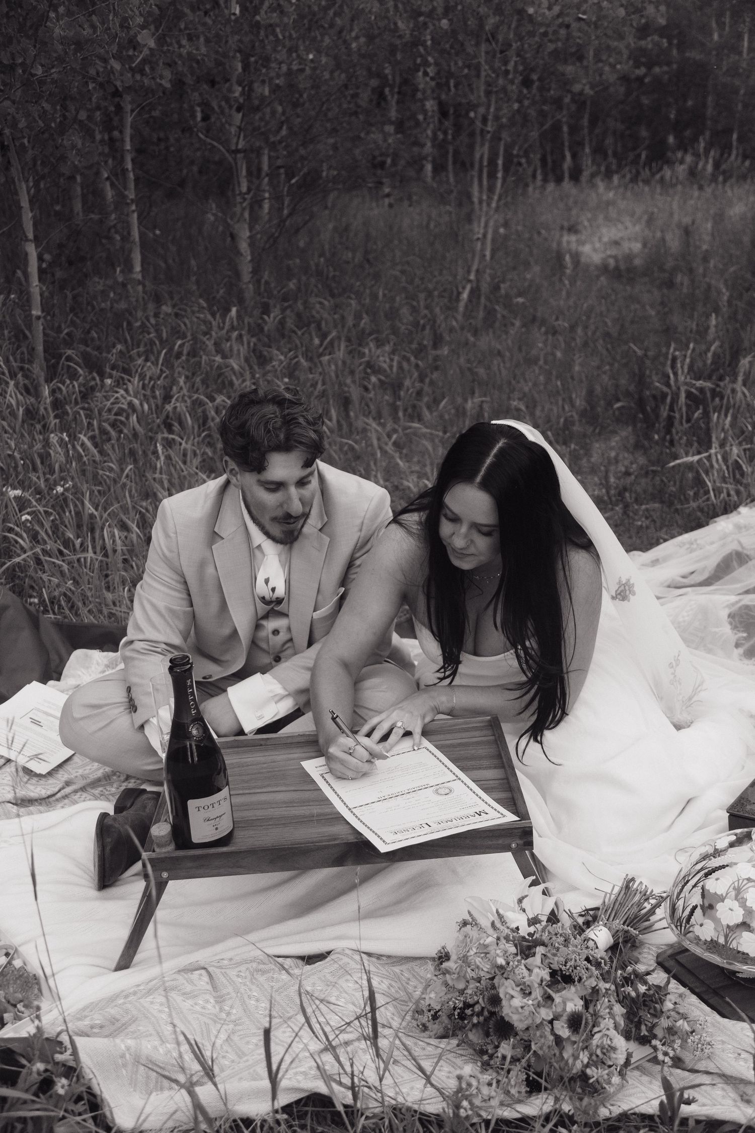 Black and white photo of newlyweds signing marriage certificate on picnic blanket with champagne.
