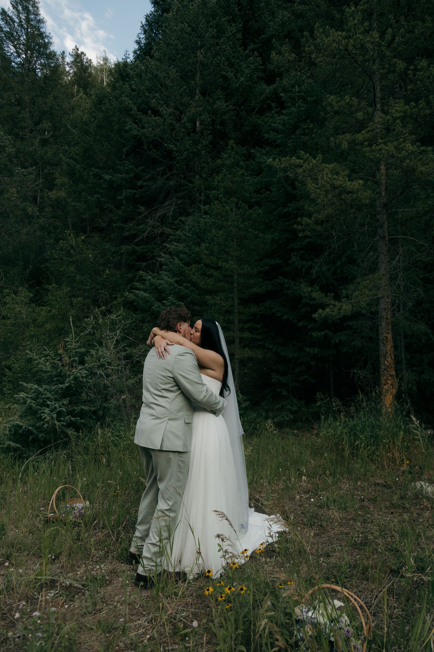 Bride and groom share a warm embrace surrounded by lush evergreen trees in a forest setting.