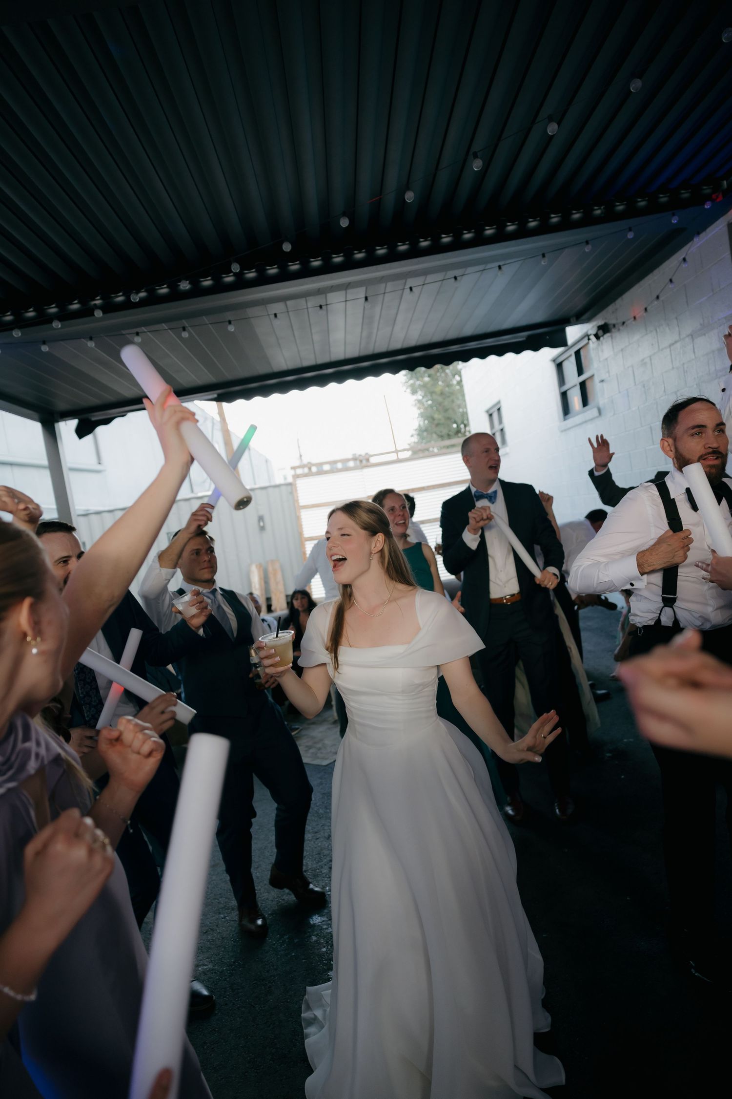 Bride walks through cheering wedding guests waving glow sticks during festive send-off celebration.