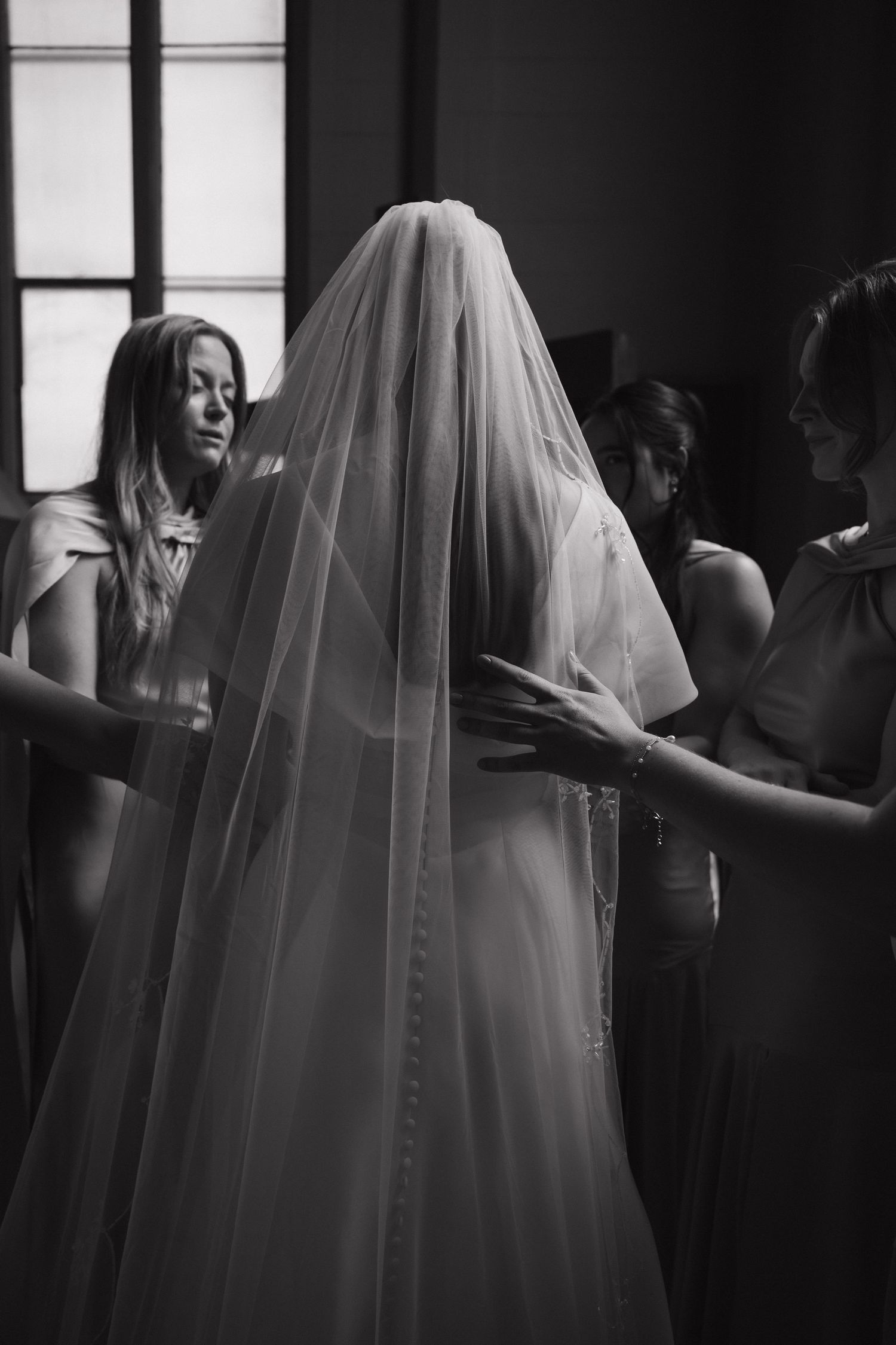 Black and white photo of bride in veil being helped by bridesmaids before wedding ceremony.