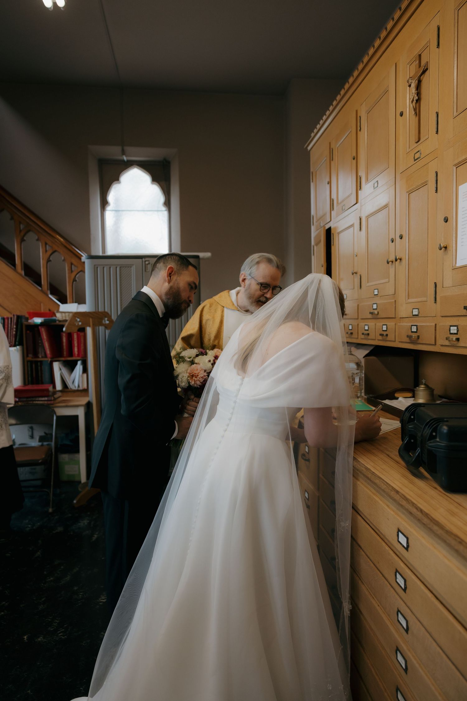 Bride and groom signing marriage documents with officiant in a church vestry.