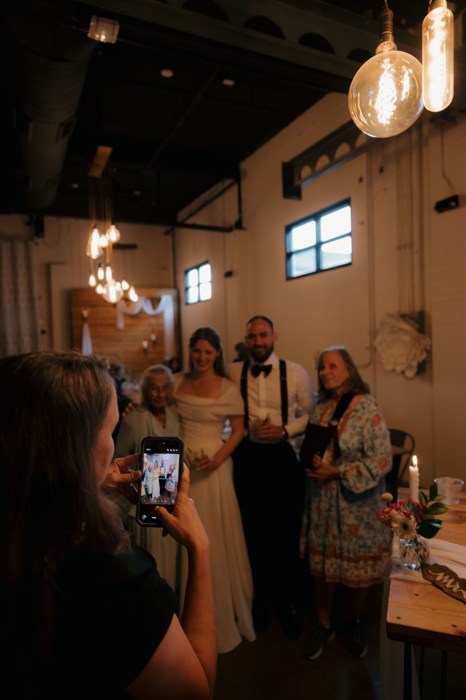 Guest photographs bride and groom posing with family members inside warmly lit wedding reception venue.