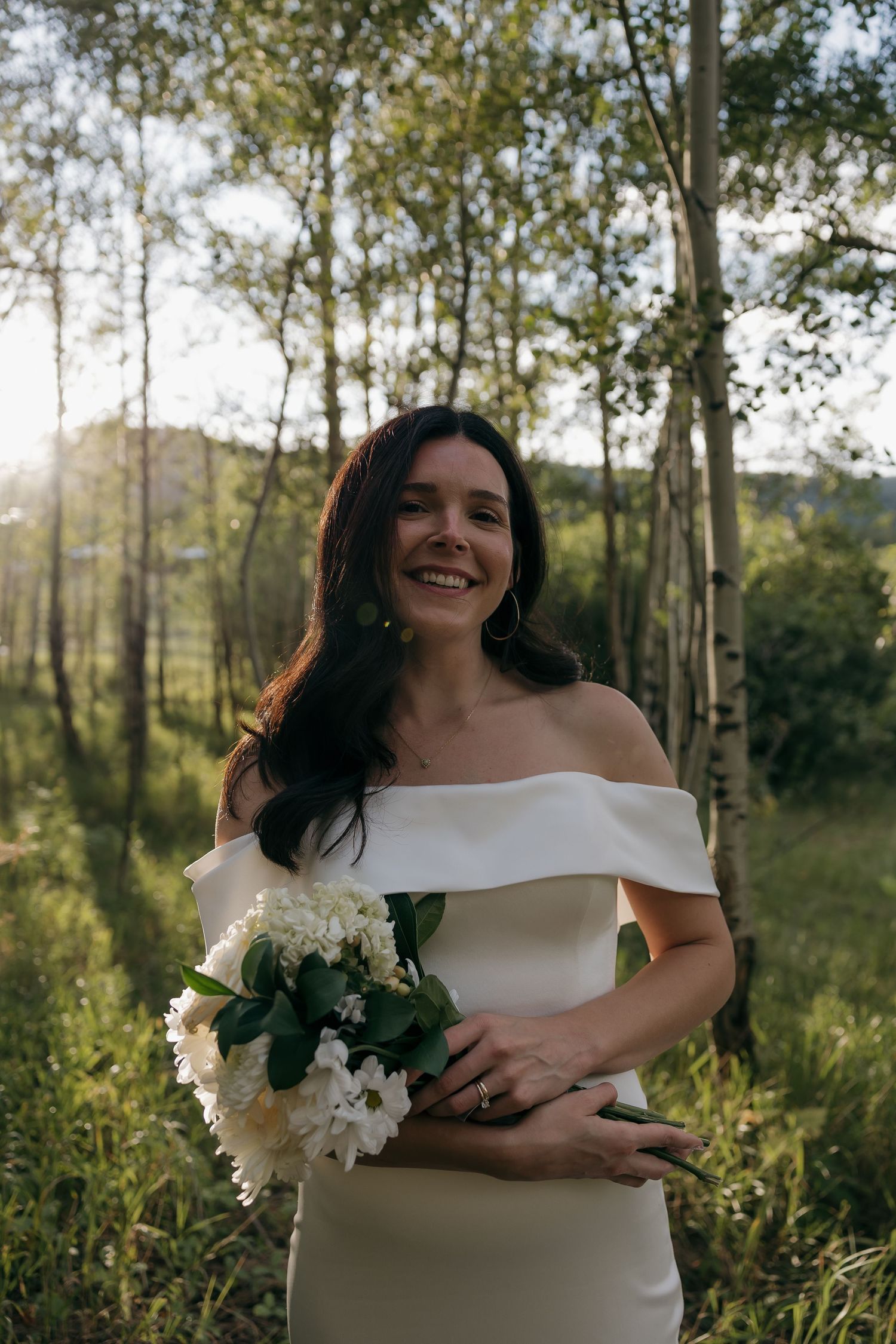 Bride in off-shoulder white gown holding white floral bouquet smiling in sunlit aspen forest.
