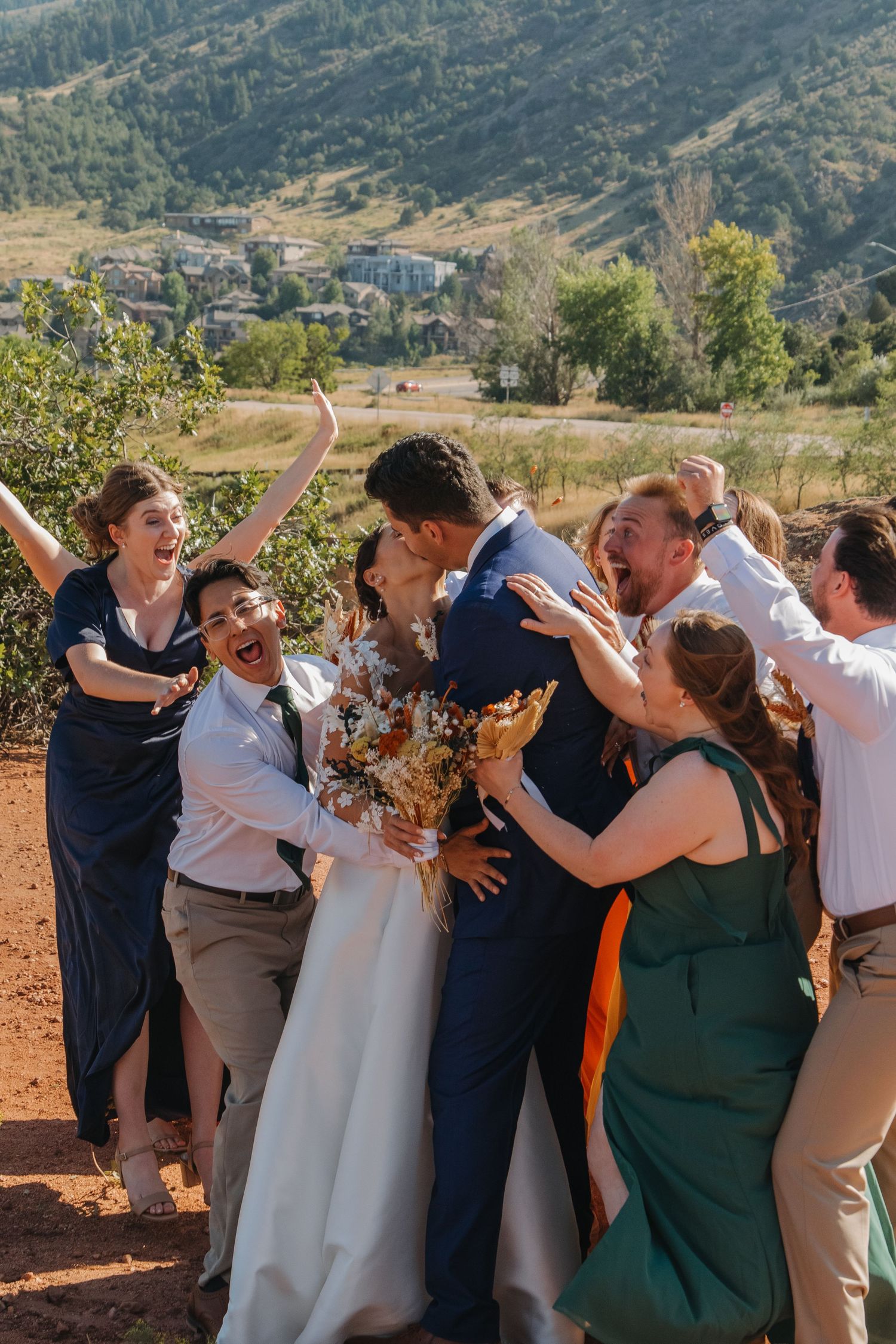 Candid moment of wedding party laughing and embracing outdoors with mountains in background.