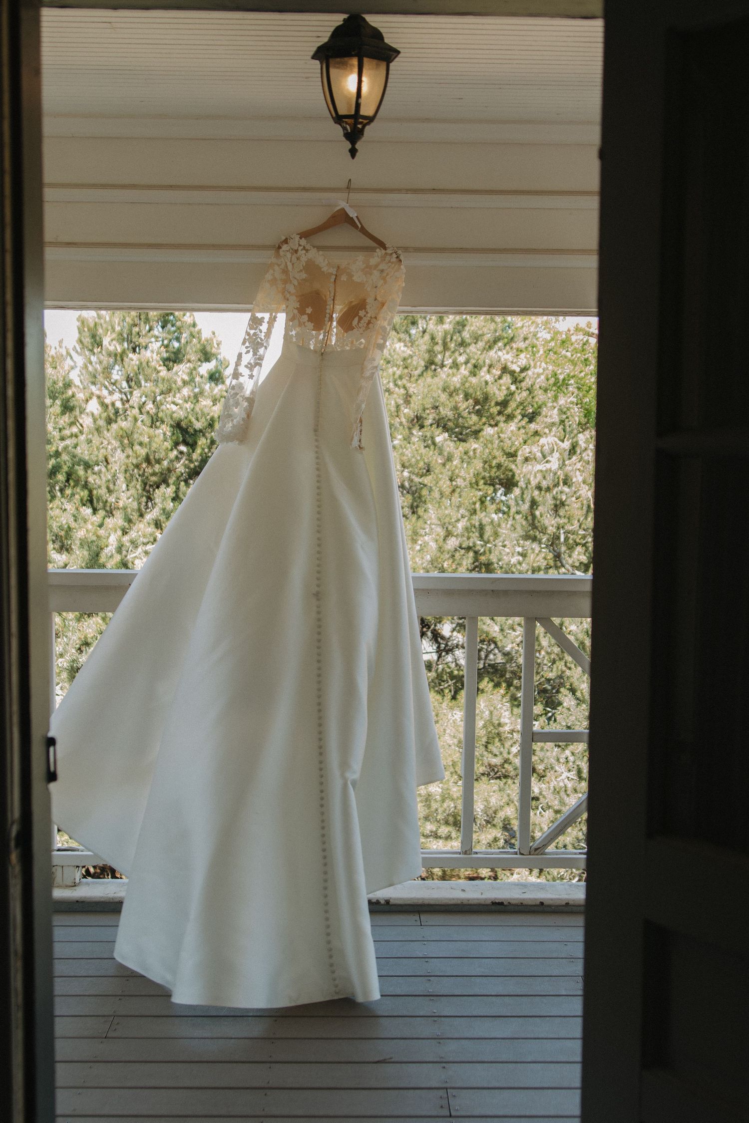 White wedding dress displayed against sunlit windows on a porch with trees visible in the background.