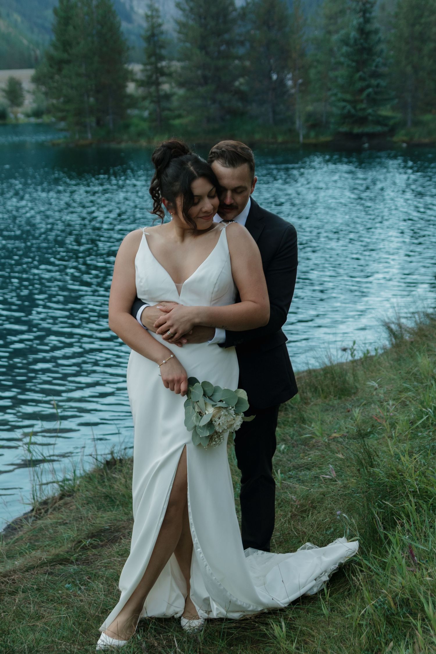 A couple embraces on a grassy shore with a mountain lake and pine forest backdrop.