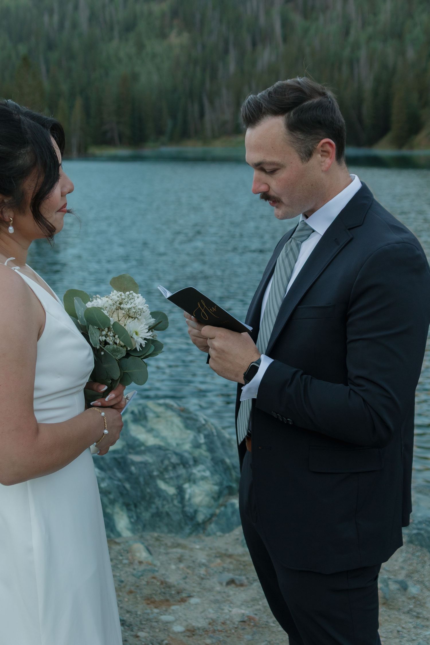 A couple in wedding attire exchanges vows by a serene mountain lake in an outdoor ceremony.