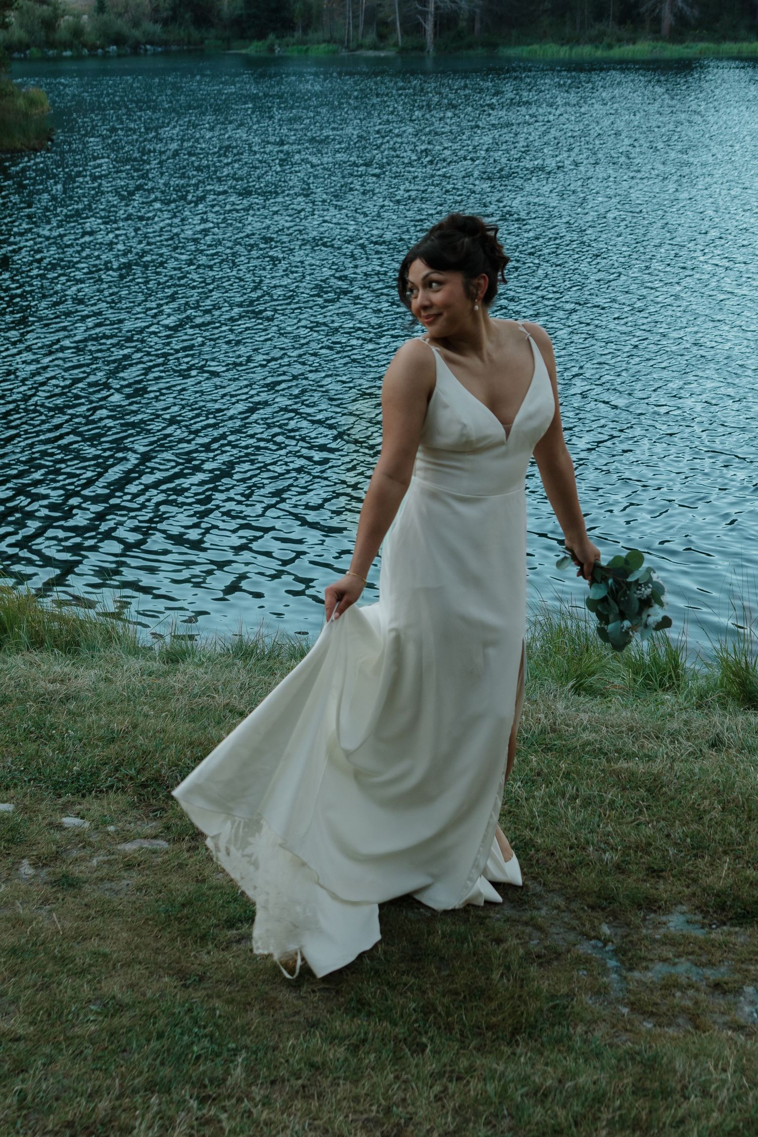 A bride in white dress twirls playfully by a serene mountain lake.
