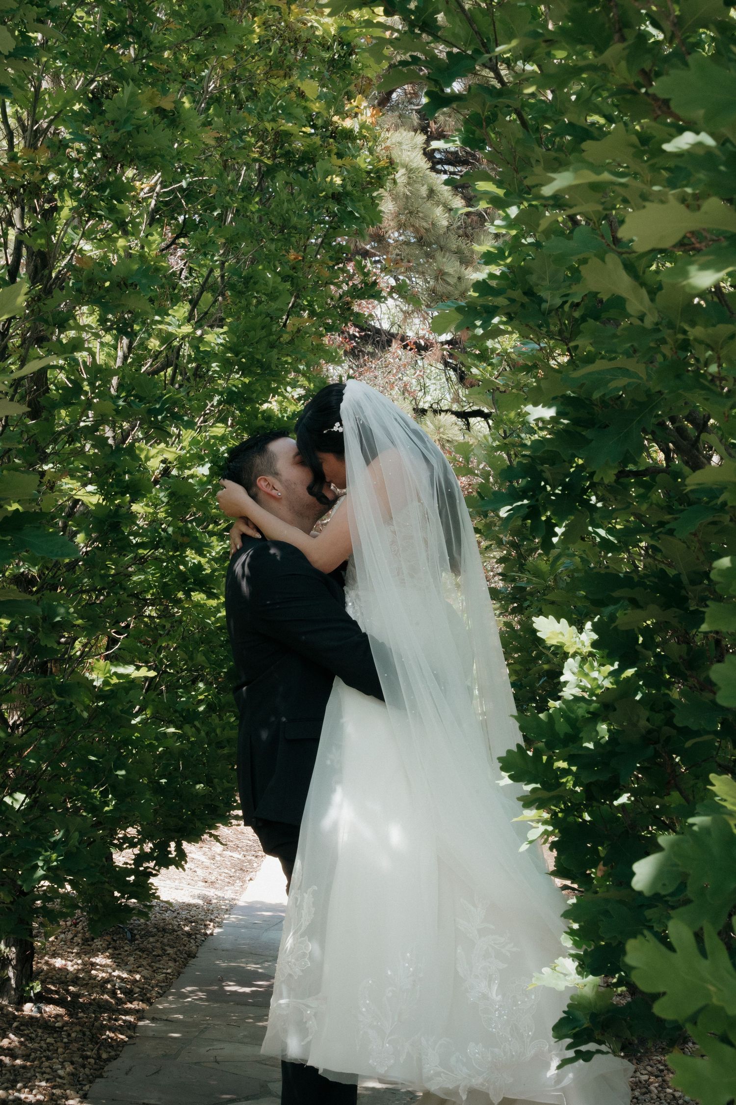 Bride and groom share a romantic kiss surrounded by lush green trees on their wedding day.