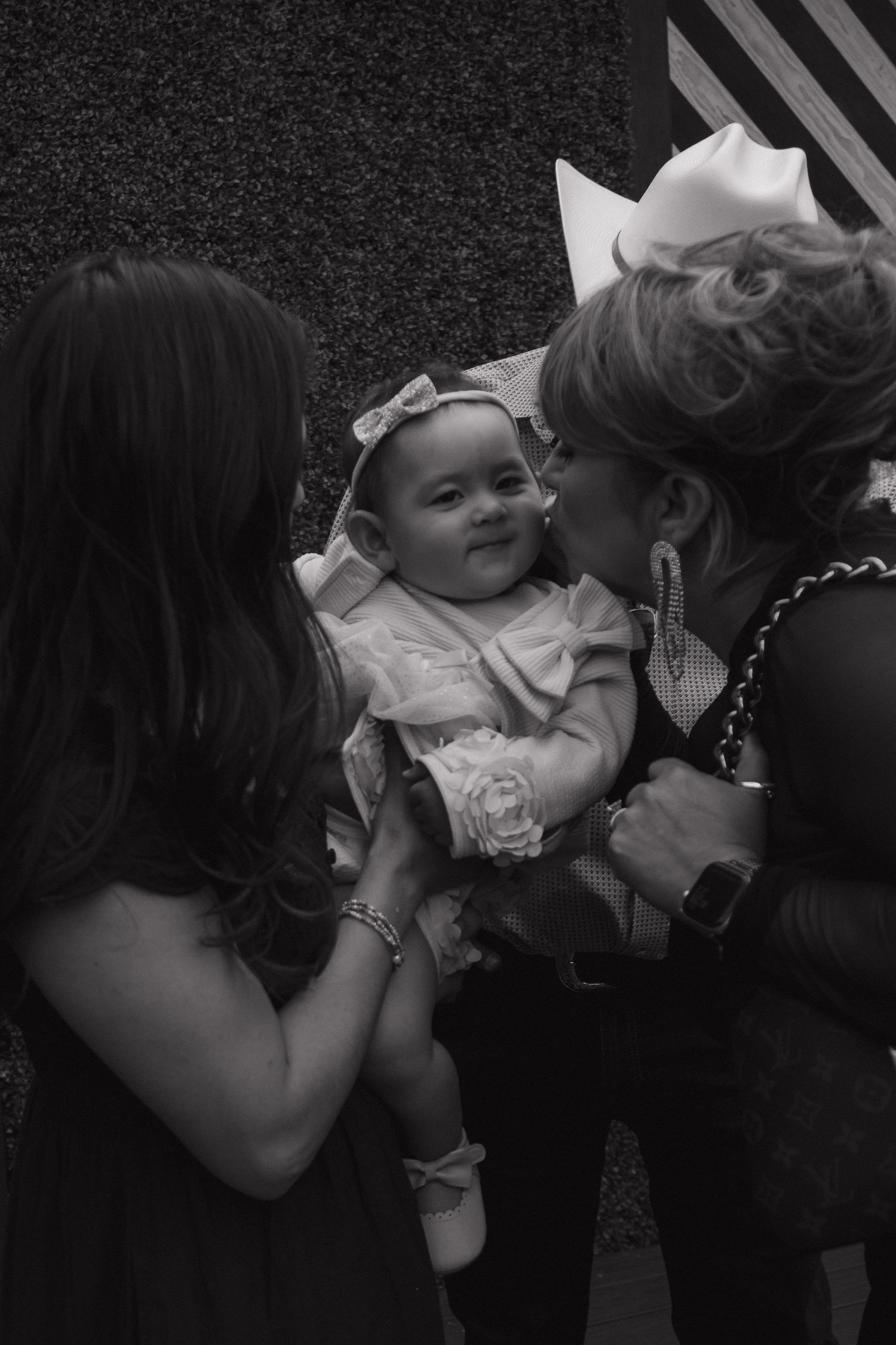 Grandmother kissing smiling baby girl in white dress at family celebration in black and white.
