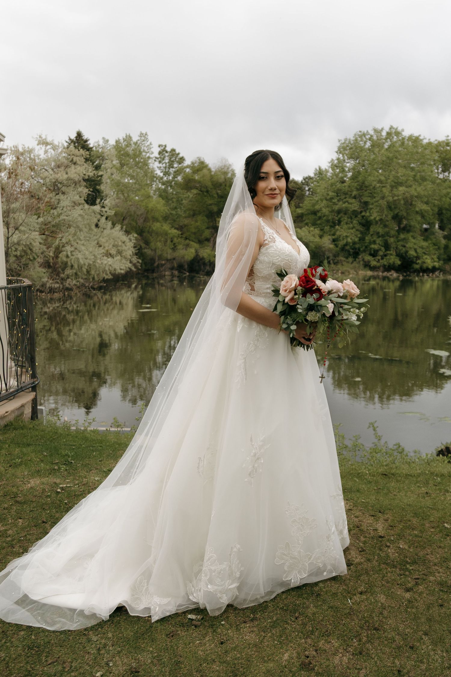 Elegant bride in lace ball gown and veil holding red and blush bouquet beside a serene pond.
