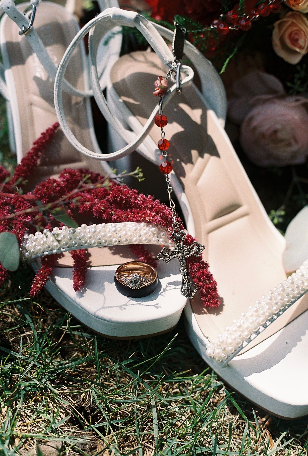 White pearl bridal heels with ankle straps, gold rings, and red crystal jewelry on green grass.