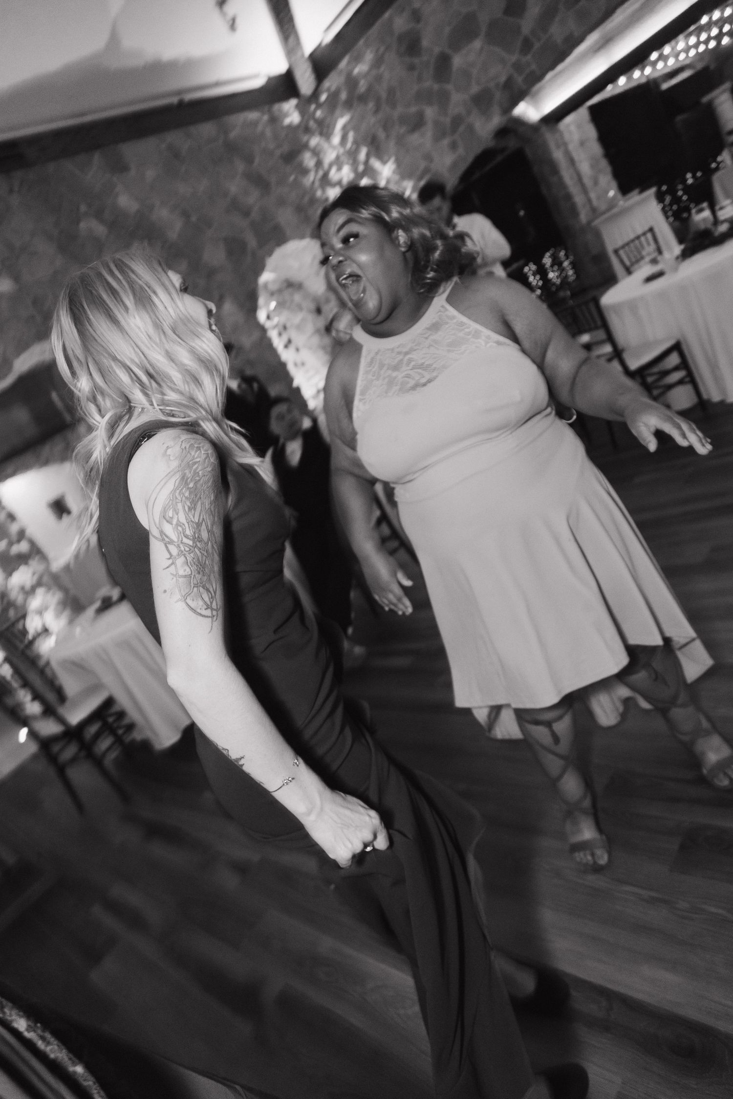 Two female wedding guests dance energetically together at reception in black and white photo.