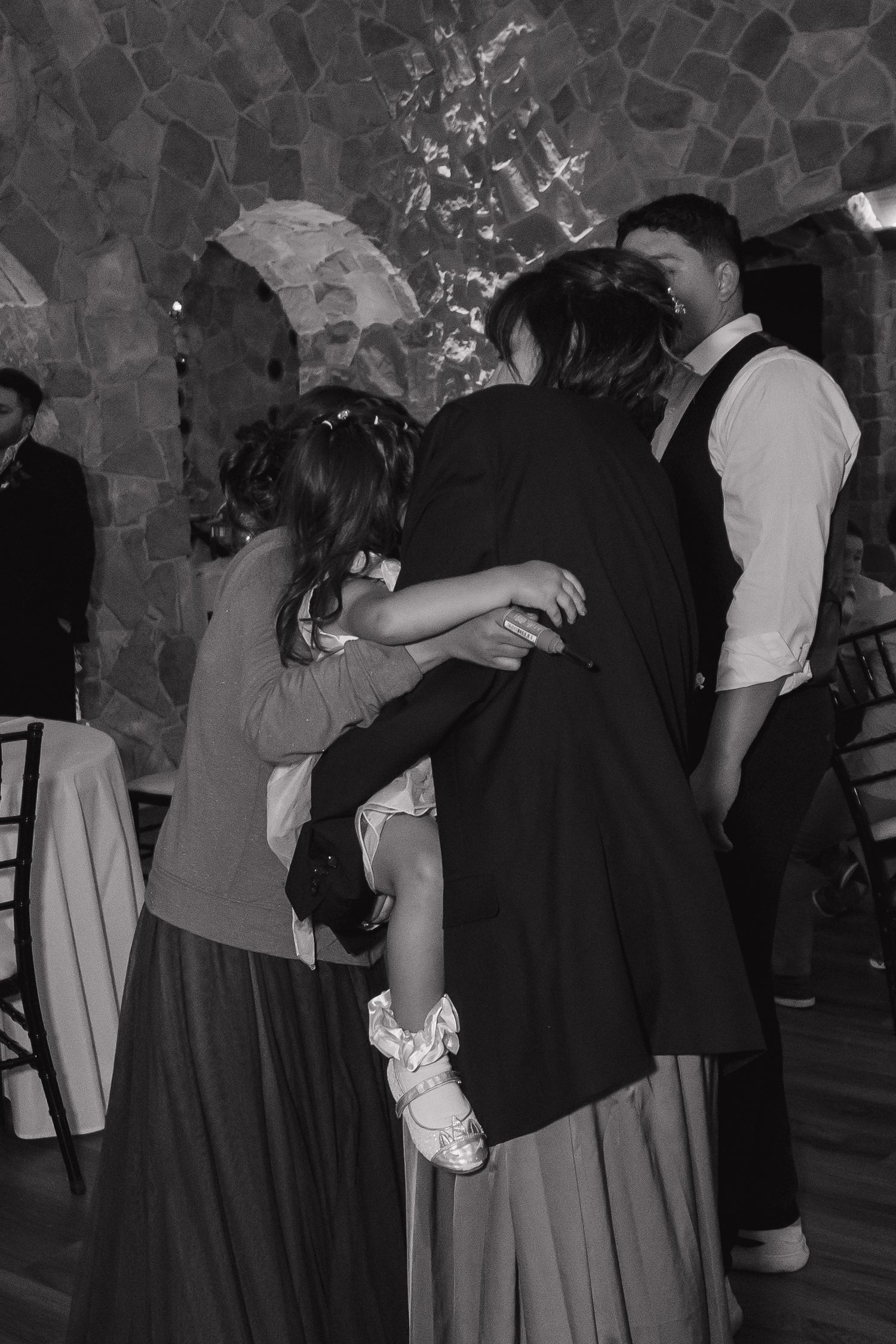 Guests share a warm group hug on the dance floor at a wedding reception in a stone-walled venue.