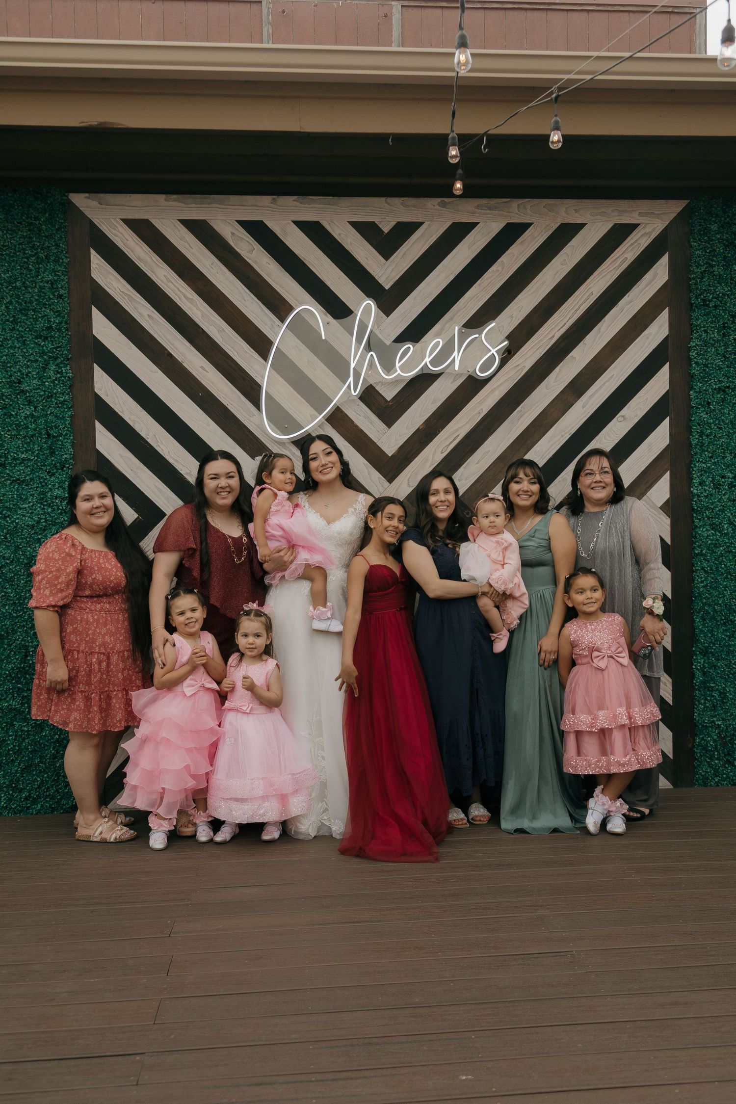 Bride in white gown poses with family and children in front of striped Cheers wedding backdrop.